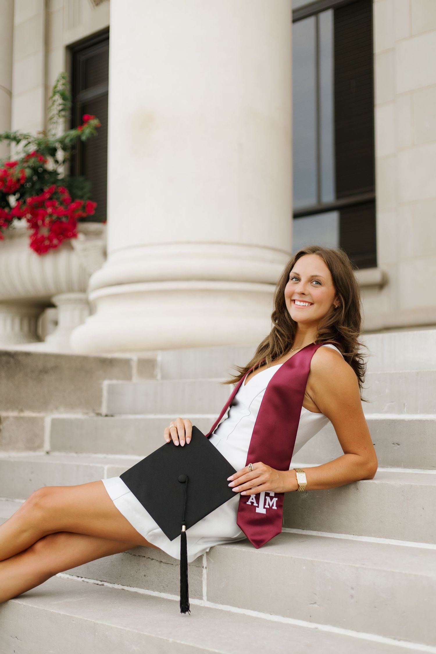 Graduate in maroon stole poses with diploma on white stone steps of academic building.