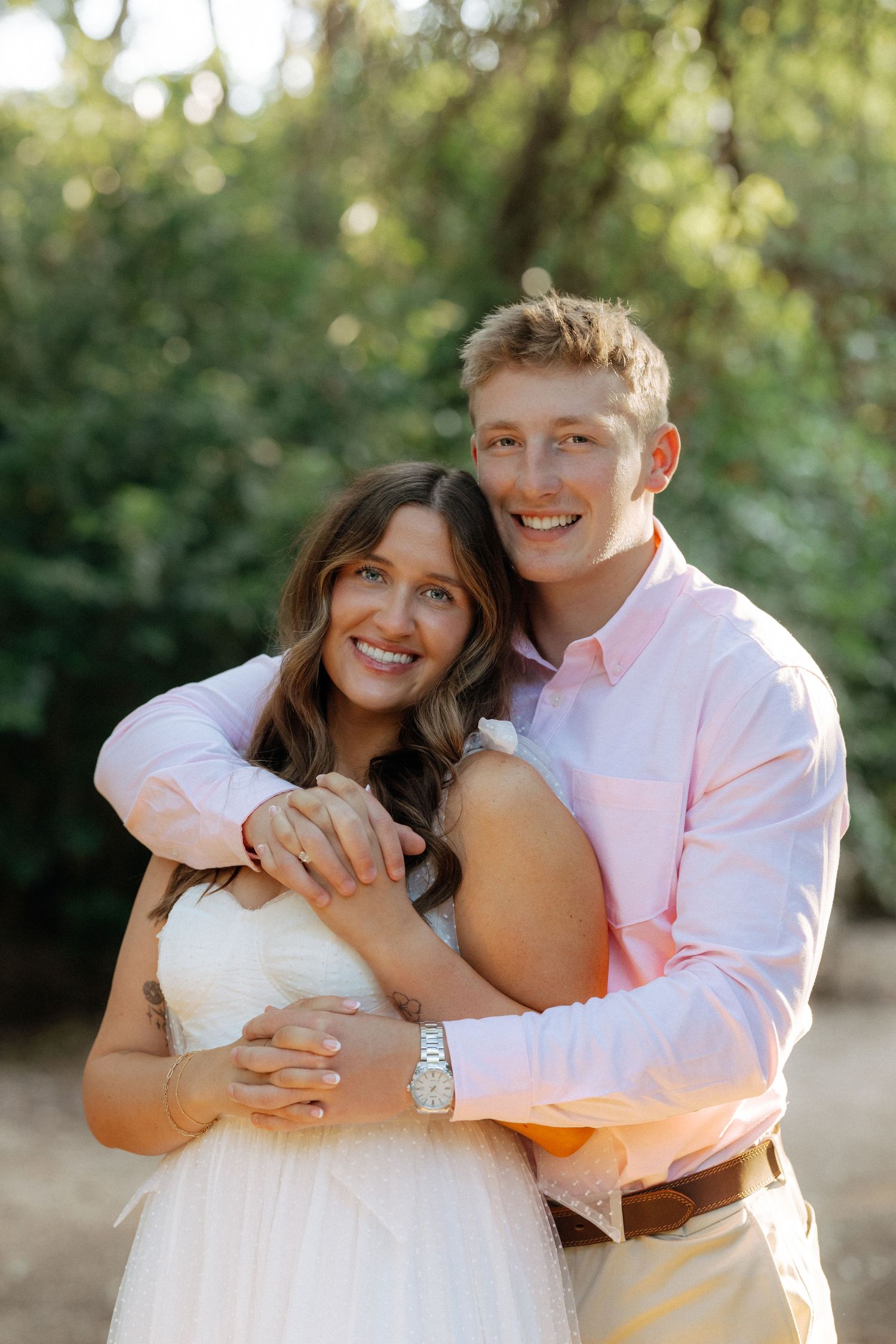Happy couple embracing outdoors, woman in white dress and man in pink shirt, smiling in a lush green setting.
