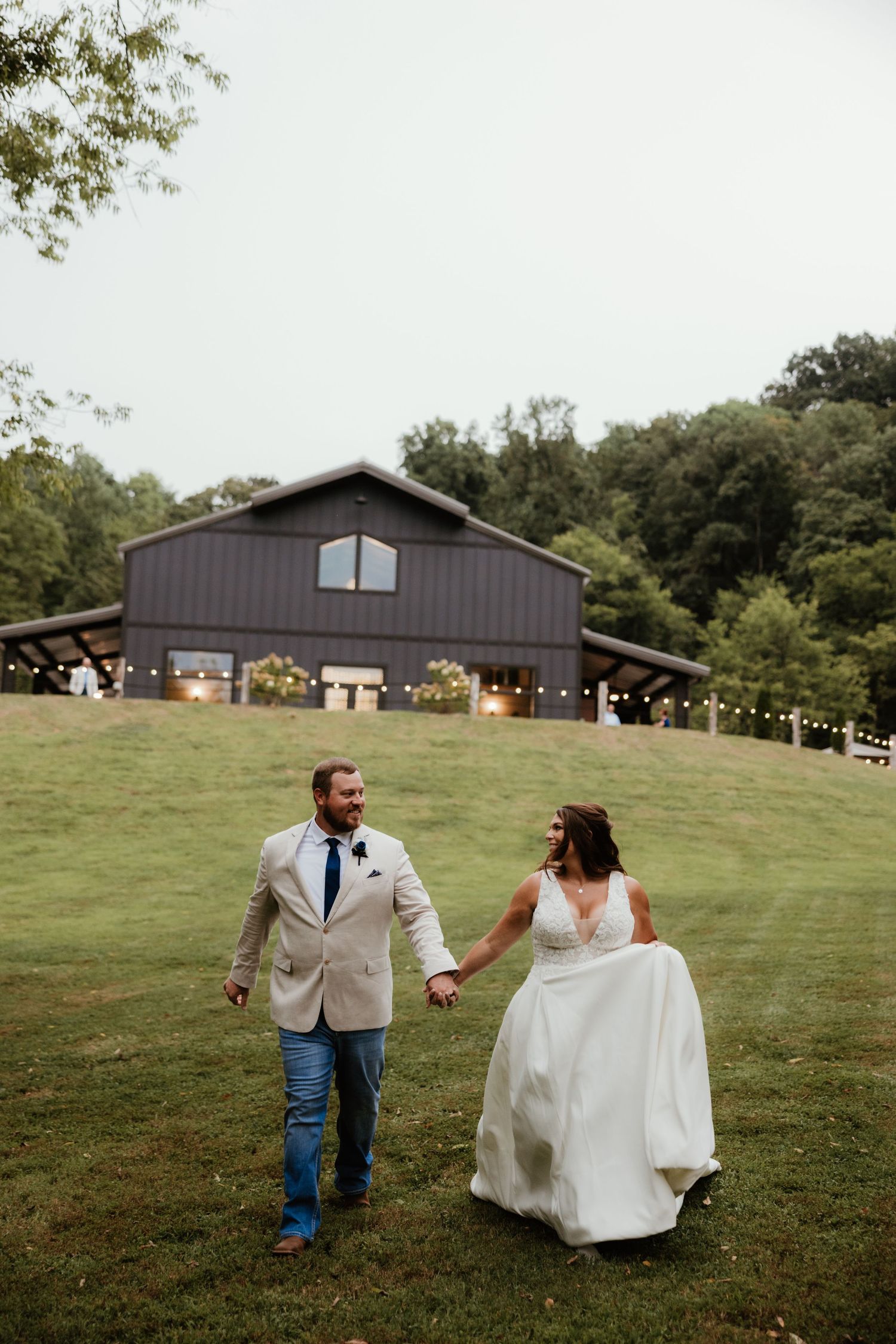 Newlyweds dance and twirl on a grassy hill with a rustic barn in the background.