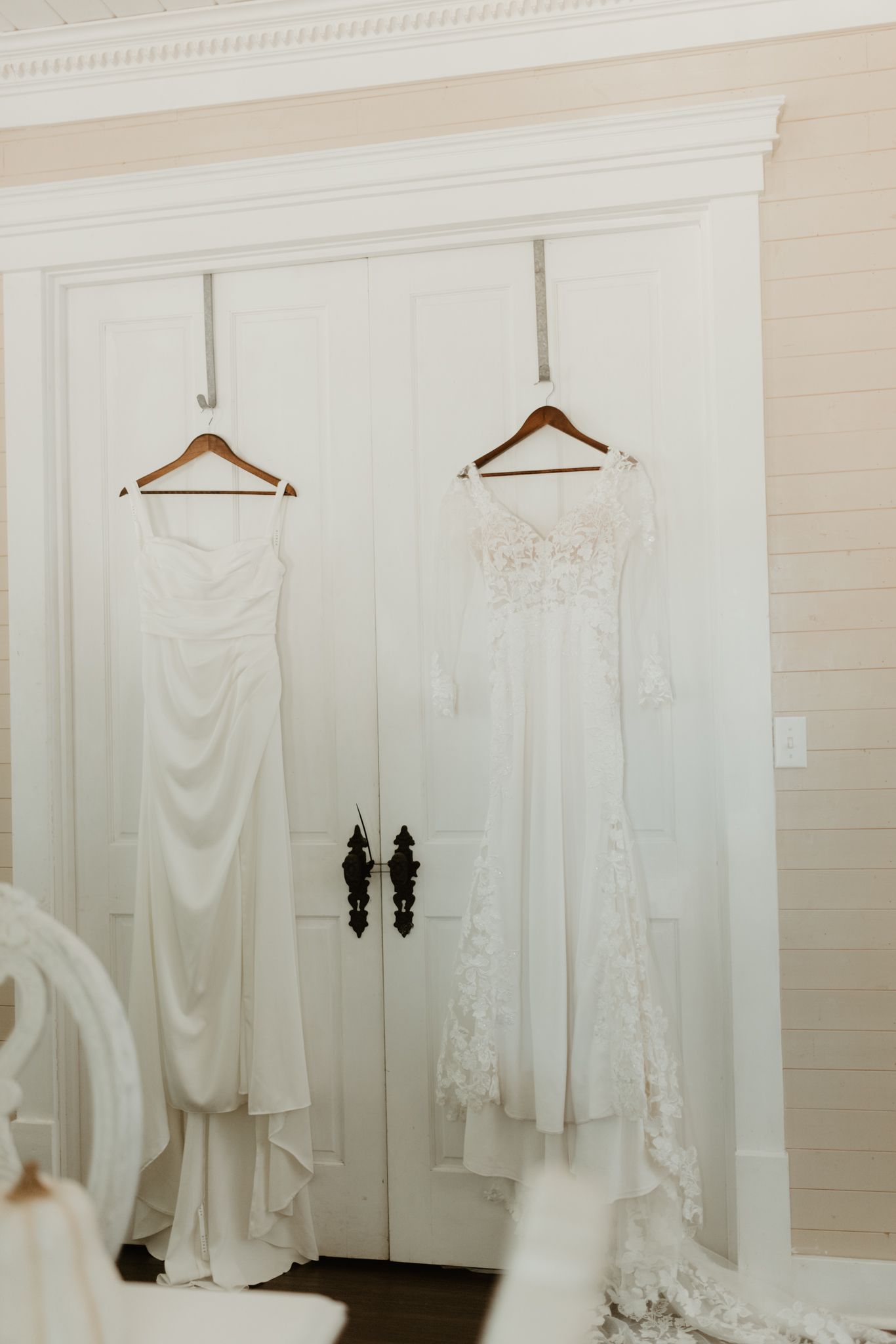 Two white wedding dresses hanging on wooden hangers against elegant white double doors.