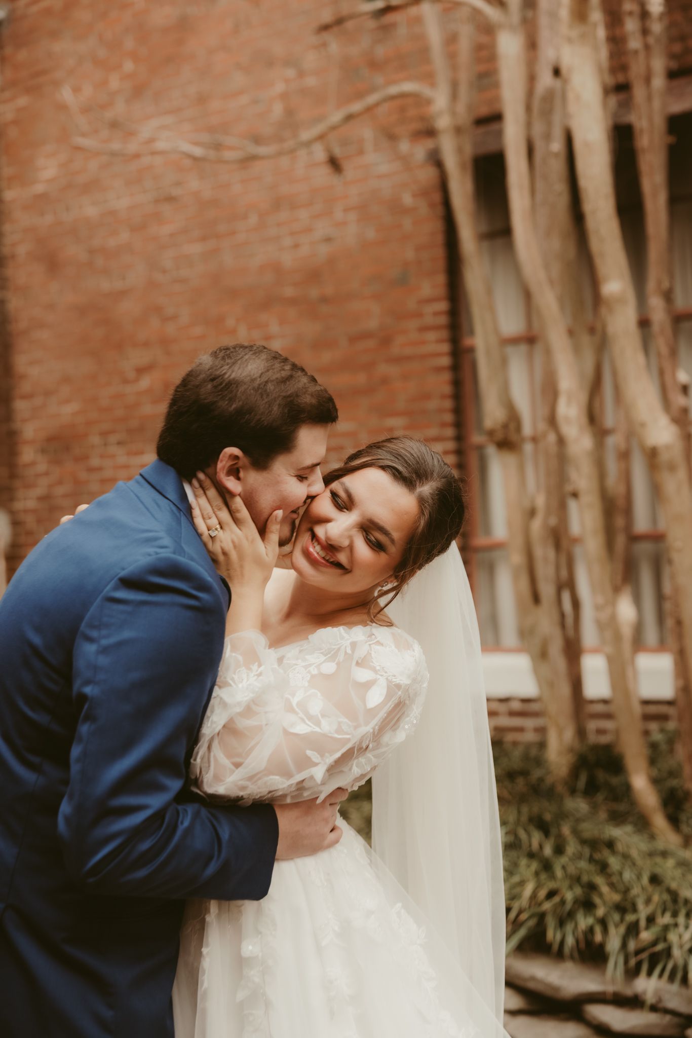 A tender moment between newlyweds captured against a weathered brick wall backdrop.
