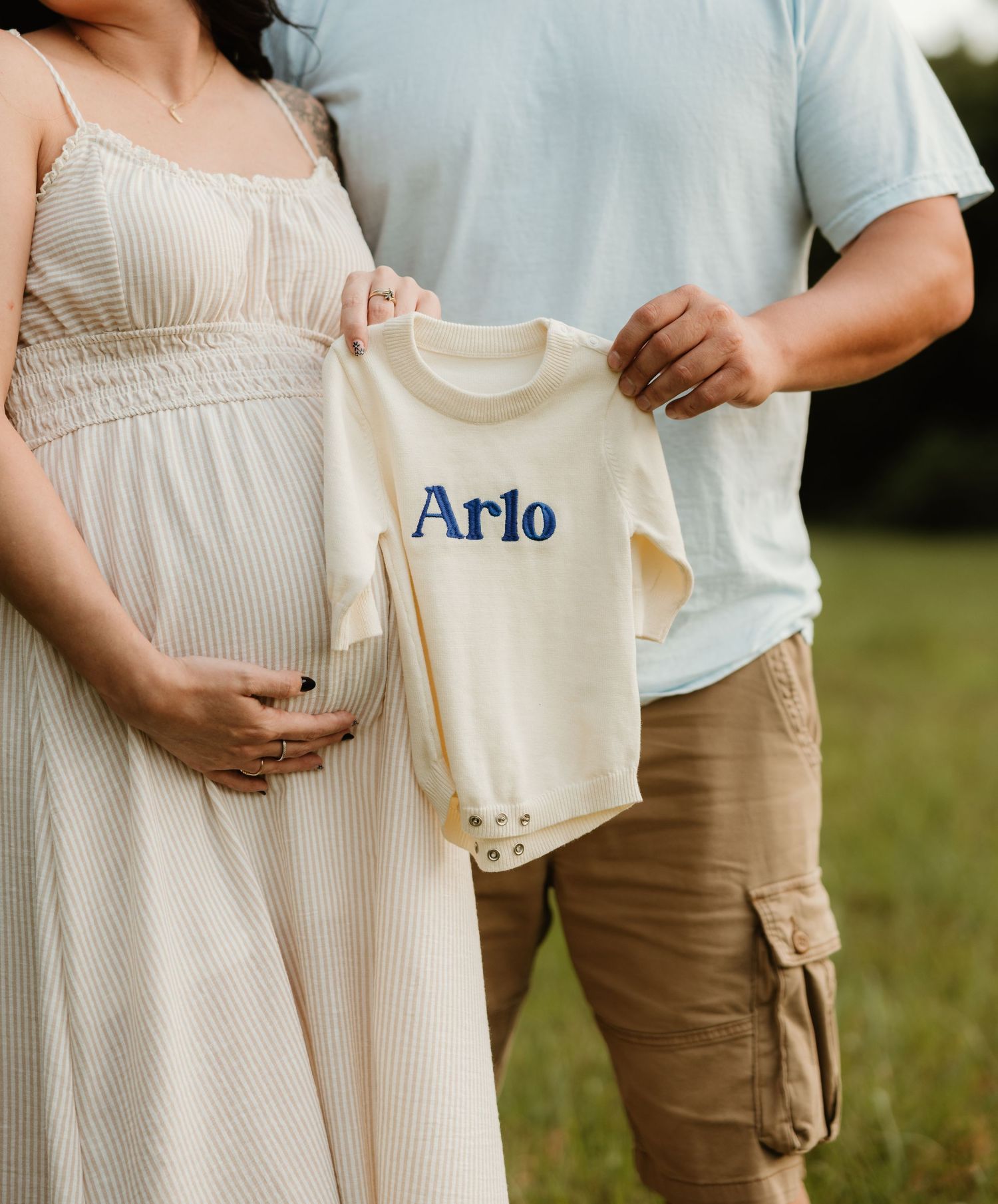 A couple holds a cream colored baby onesie with blue embroidered text spelling 'Arlo' during a pregnancy announcement.