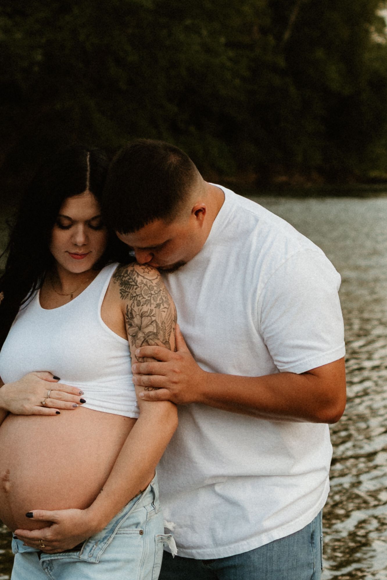Maternity photo series showing couple in white tops embracing by a lake, sharing intimate moments with growing baby bump.