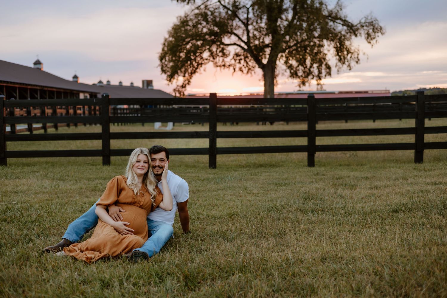 Silhouettes of figures at sunset relaxing on a grassy hillside with wooden fencing and buildings in the background.