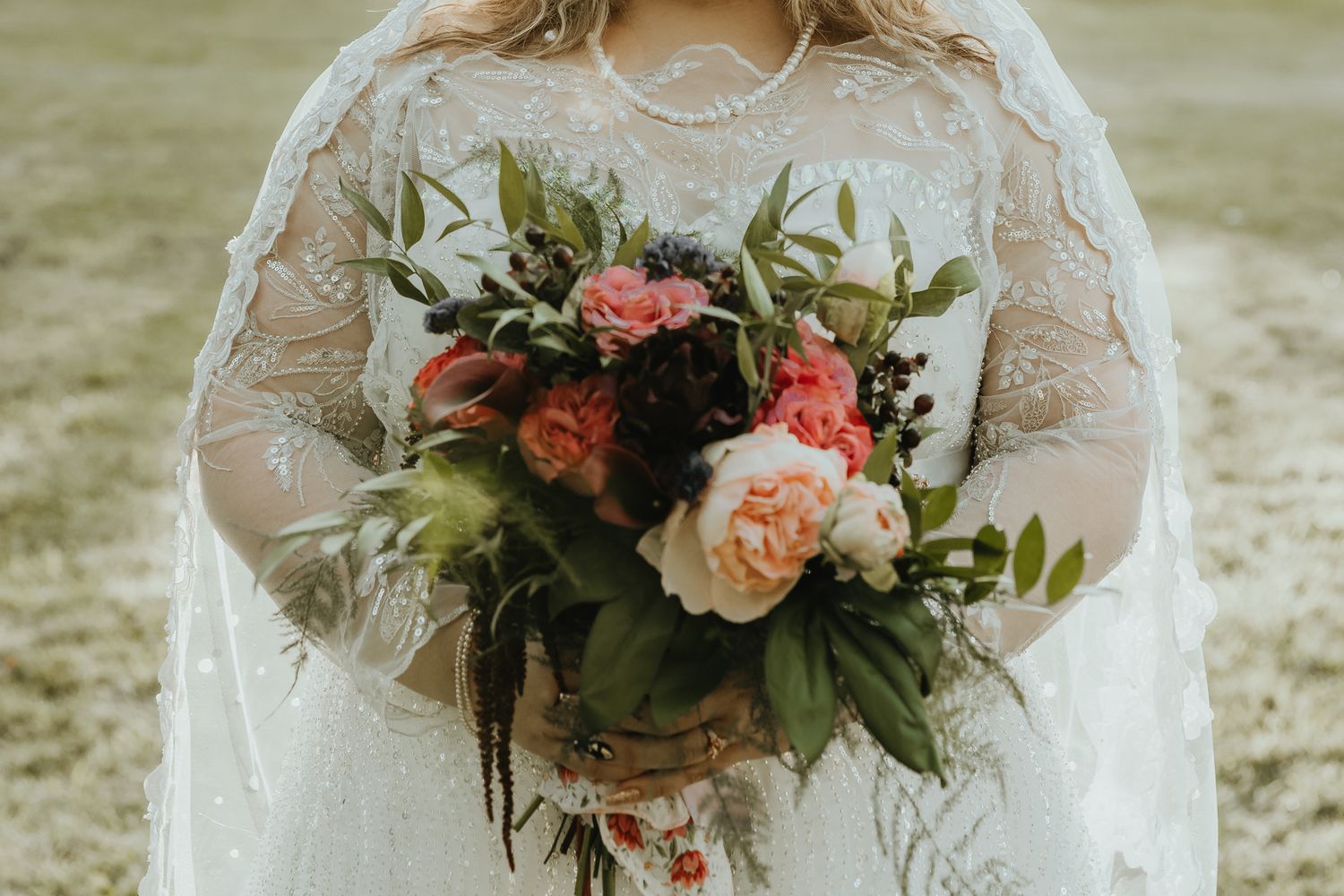 A stunning bridal bouquet of peach and coral roses with fresh greenery held against a white lace wedding dress.