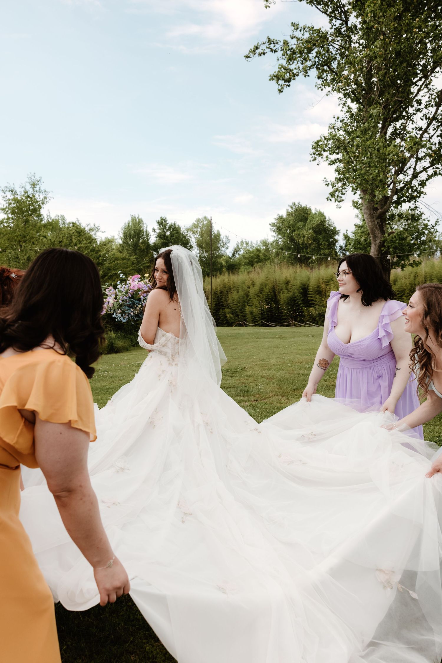 Bridesmaids in pastel dresses help arrange the flowing train of a white wedding gown on a grassy lawn outdoors.