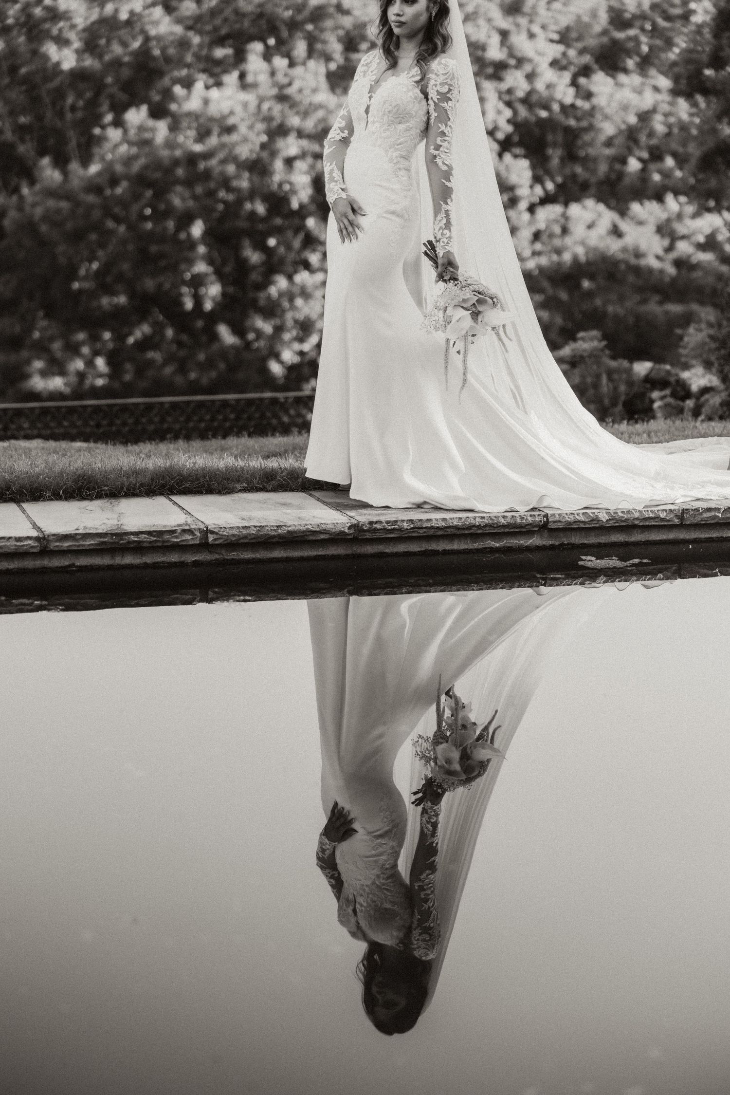 Black and white photo of a bride in a lace wedding gown with veil, reflected in still water outdoors.