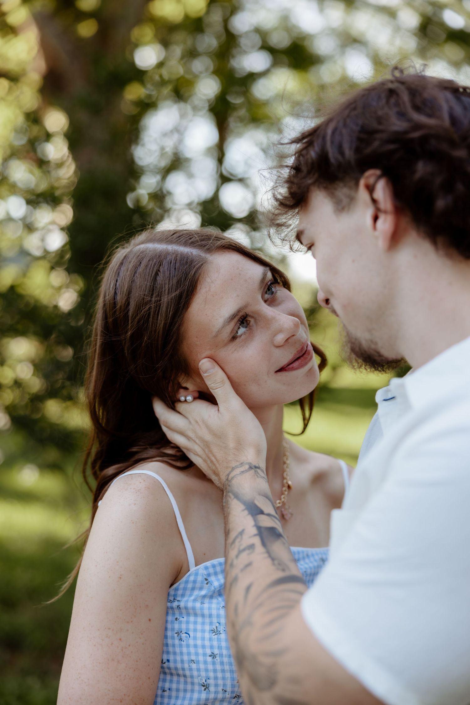 A couple shares a tender moment outdoors, man gently cupping womans face, both leaning in close surrounded by green trees.