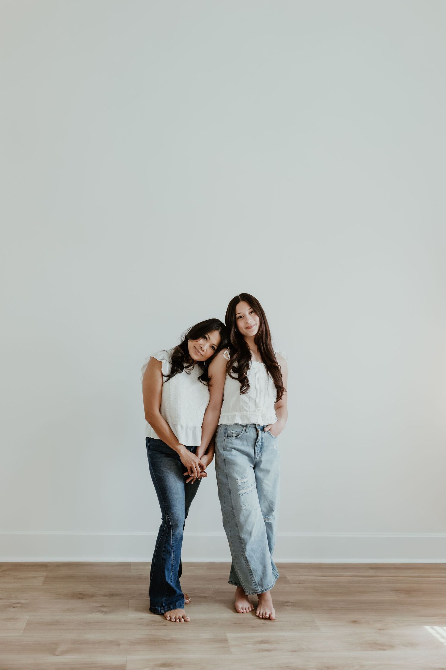Two young women in white tops and jeans pose barefoot against a light gray wall on hardwood floors.