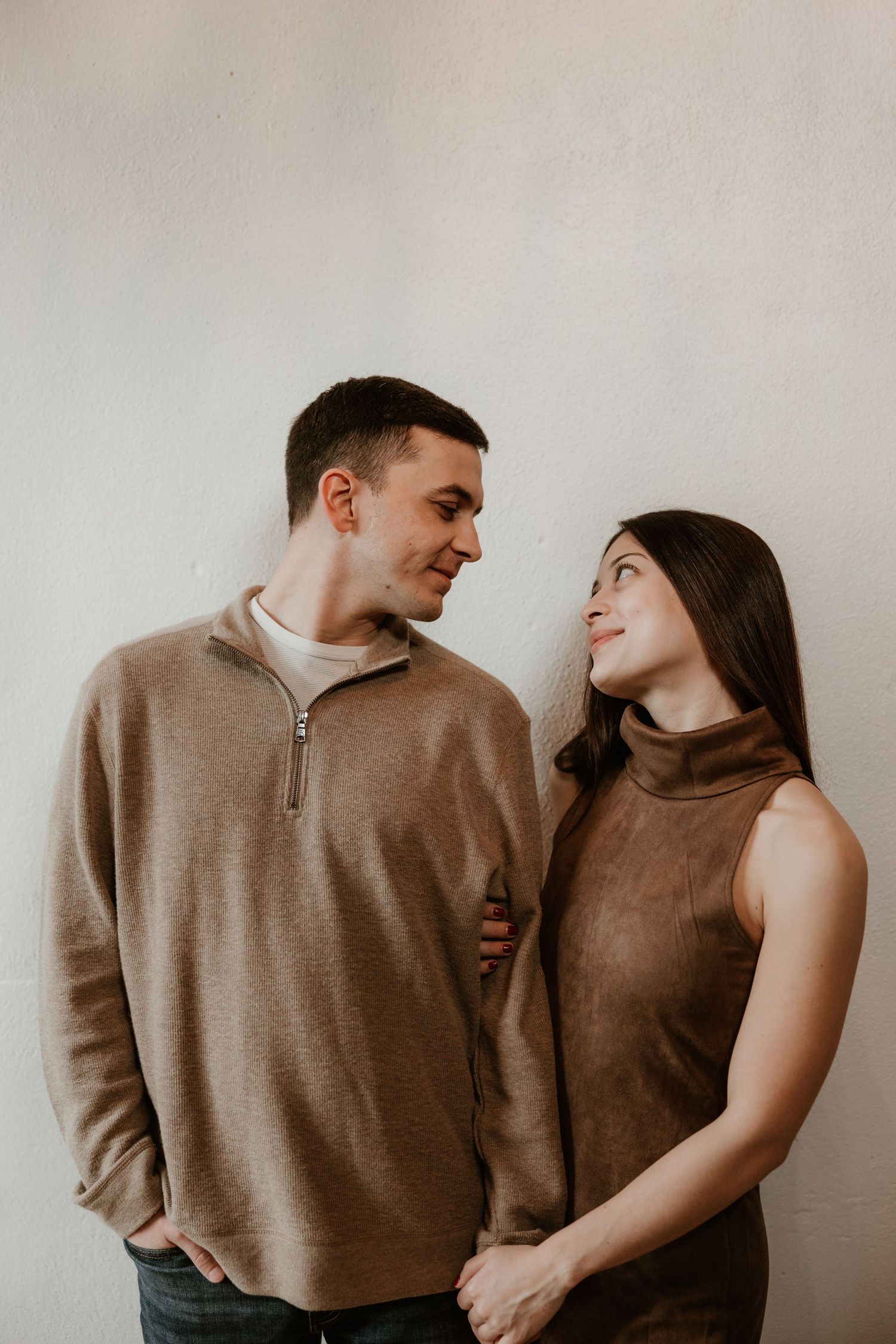 A couple in matching brown outfits smiling at each other against a white wall.