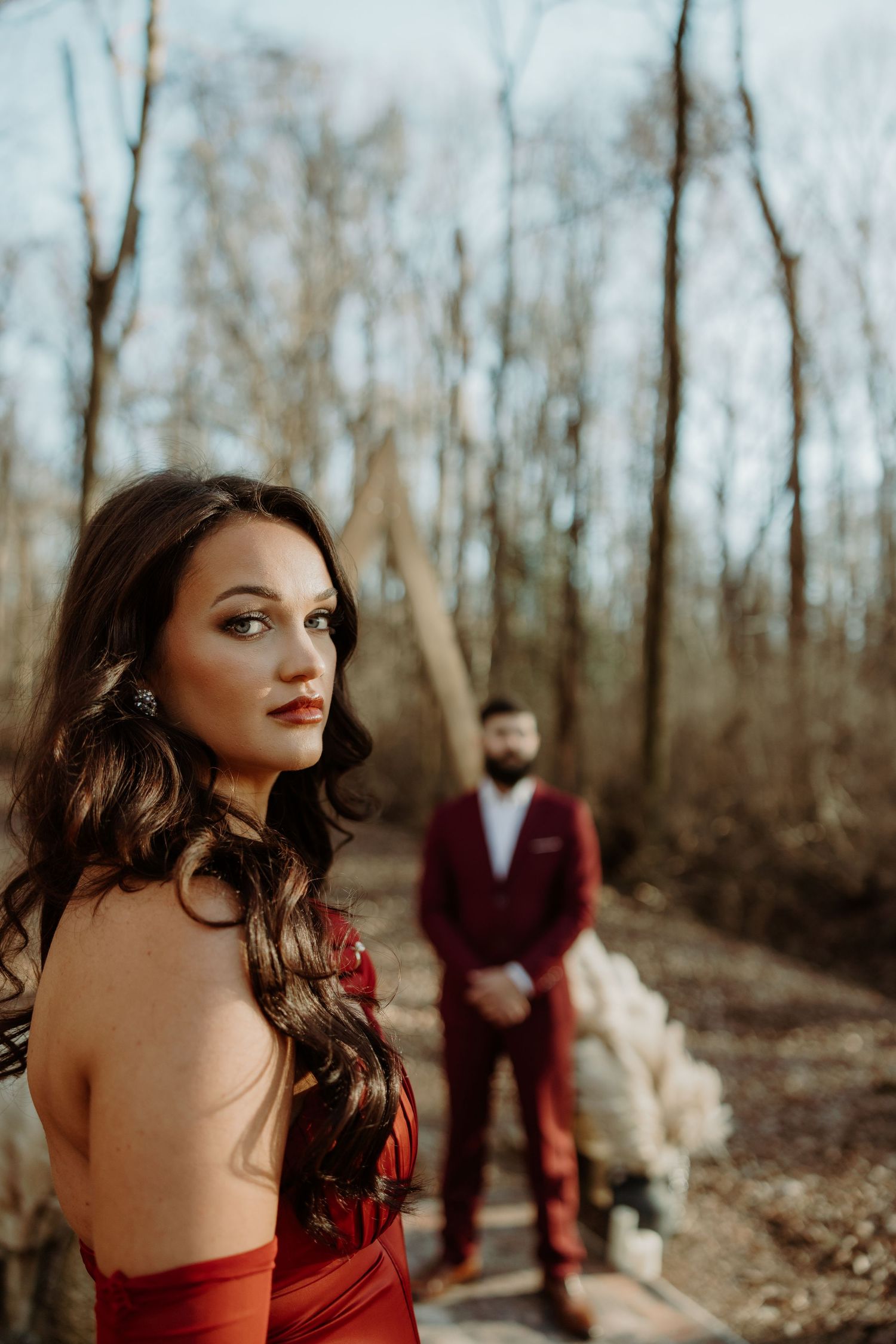 A woman in a red strapless gown poses outdoors with a bearded man in a burgundy suit standing behind her.