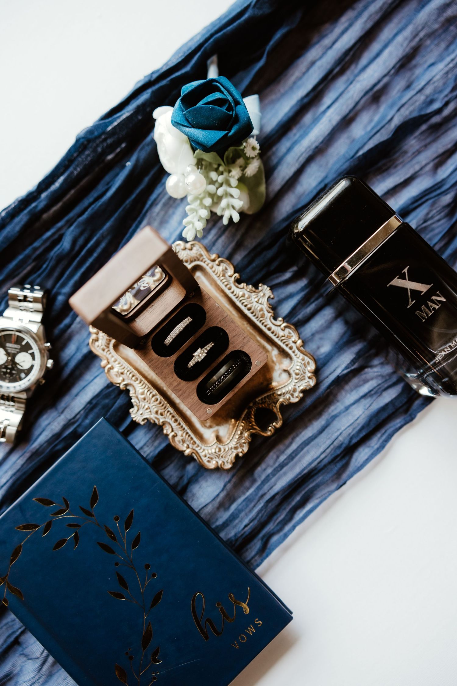 Wedding rings arranged on rustic blue wood with romantic vintage perfume bottle and delicate ivory flowers.