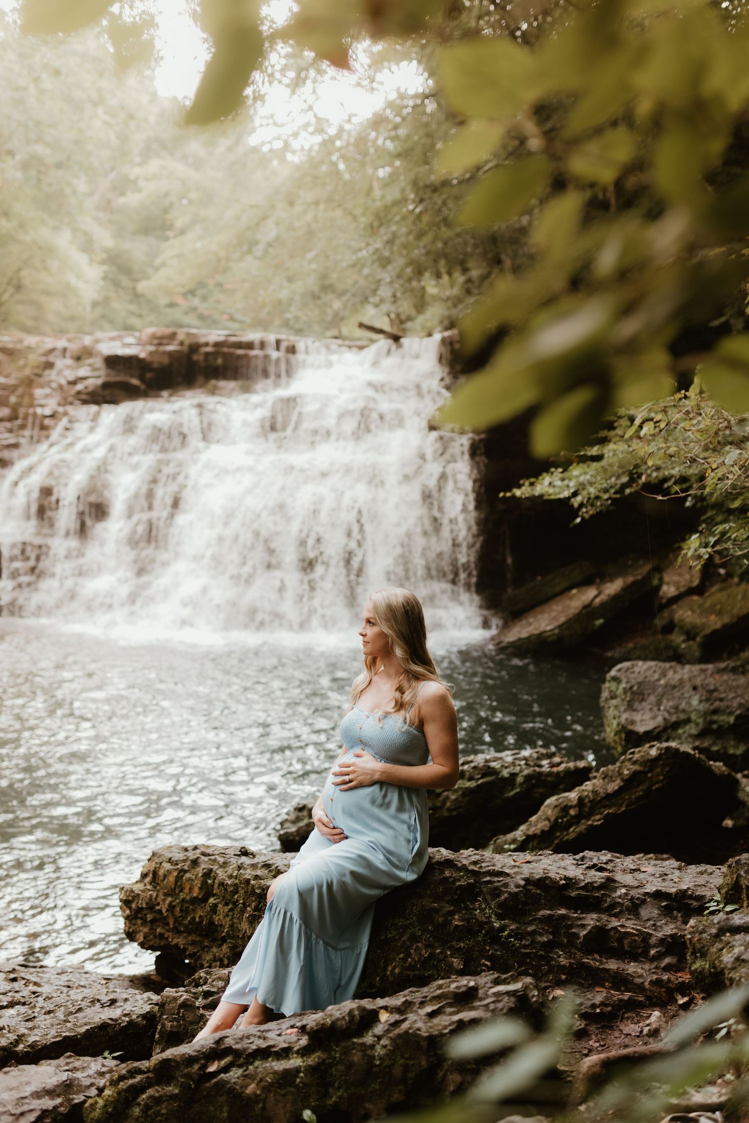 A serene scene at a waterfall where someone in a blue dress sits on rocky outcropping near cascading water.