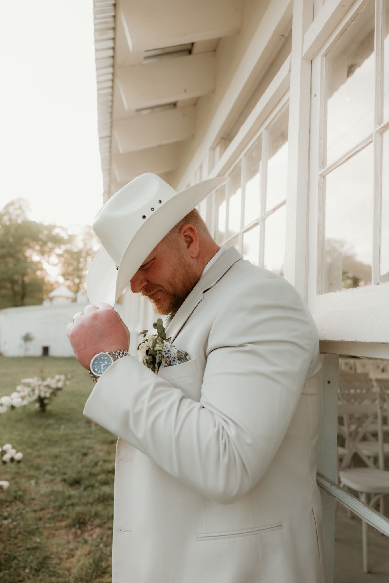 Someone wearing a white suit jacket and cowboy hat stands outside a white building in golden sunlight.
