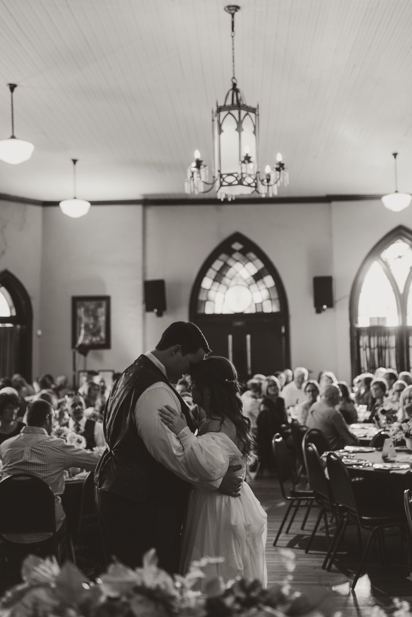Black and white photograph of a wedding reception in a church hall with vintage chandeliers and gothic windows.