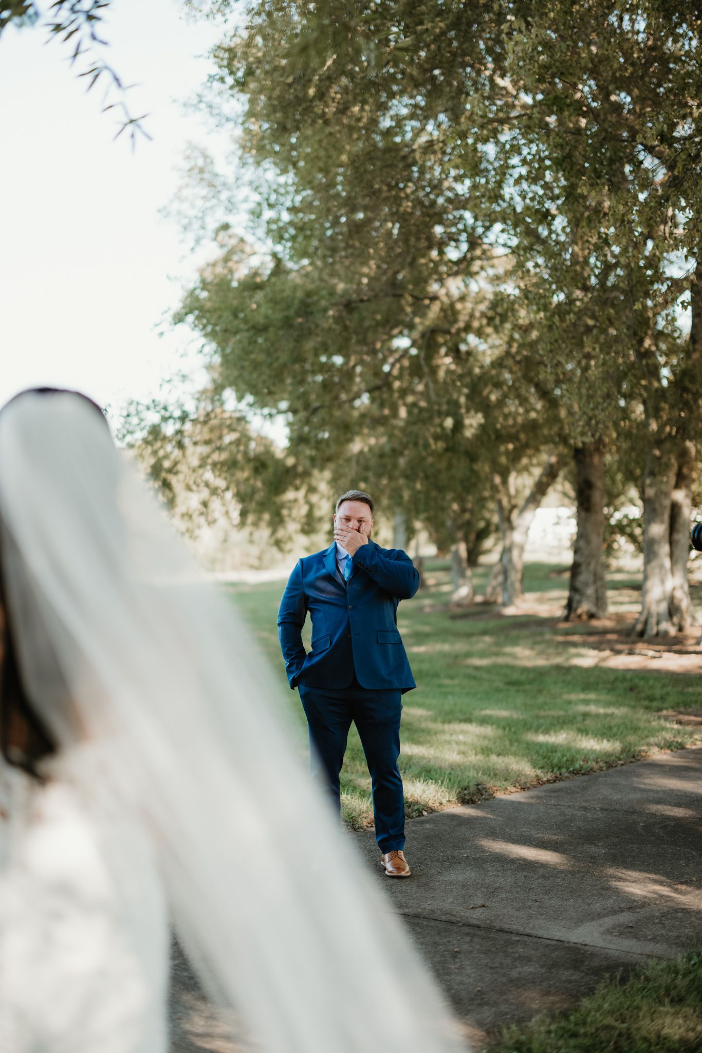 A romantic couple walks along a tree-lined path in an outdoor natural setting wearing formal attire.