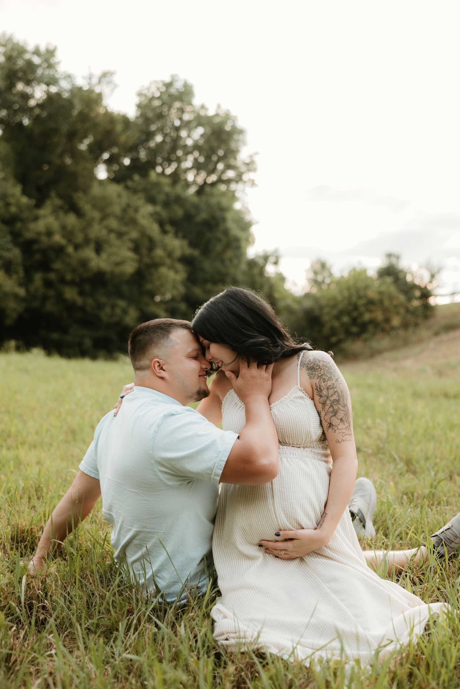 A couple in wedding attire shares intimate moments while sitting together in a grassy meadow during sunset.