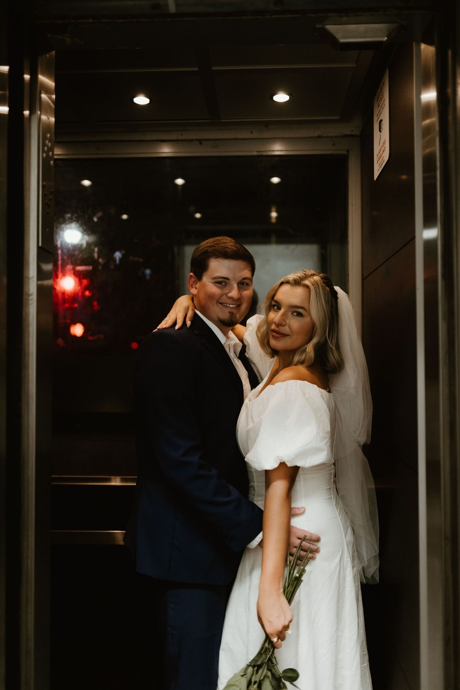 A couple poses together in a dimly lit doorway dressed in elegant wedding attire.