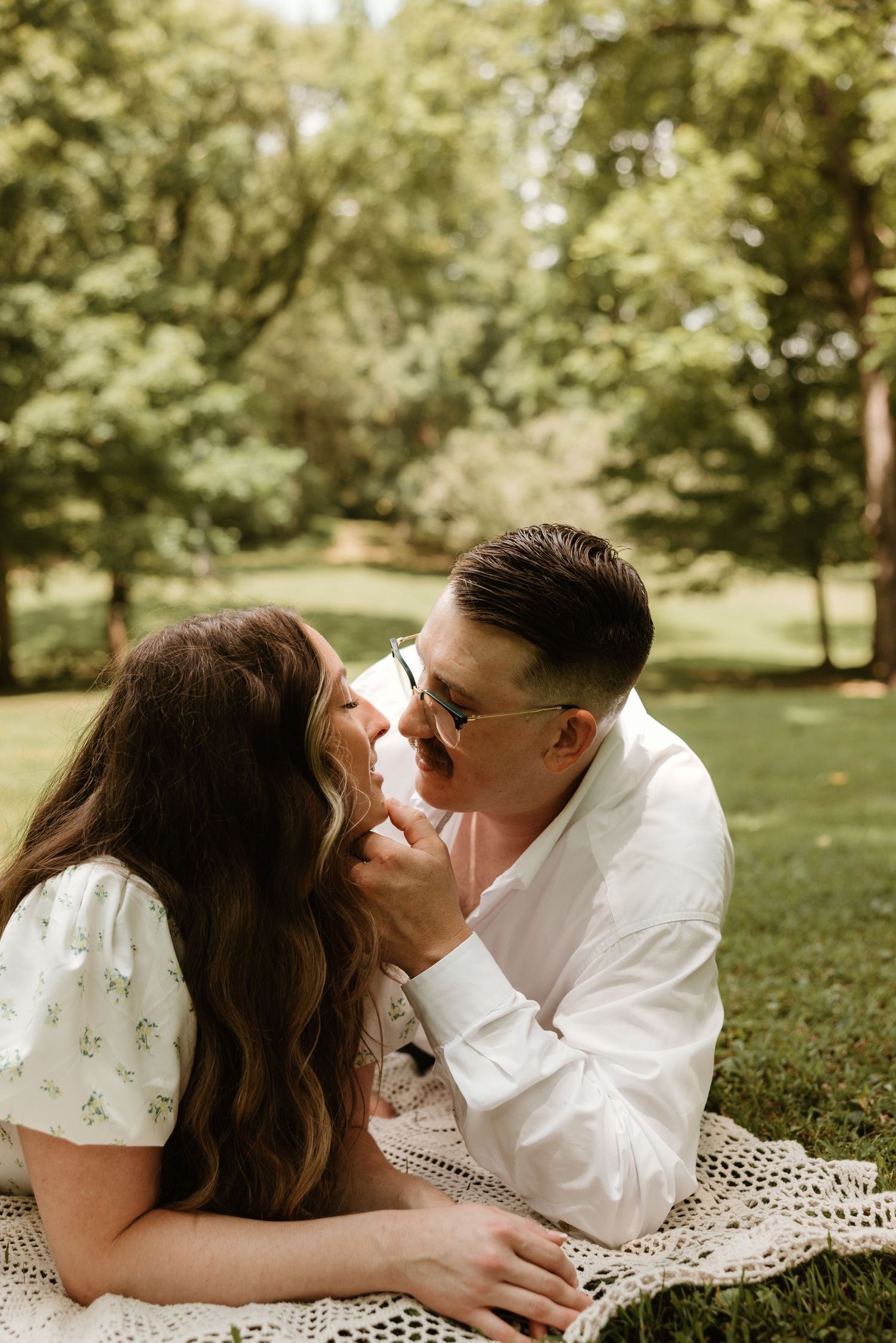 A romantic couple shares an intimate moment while laying on a blanket in a sunny park setting.