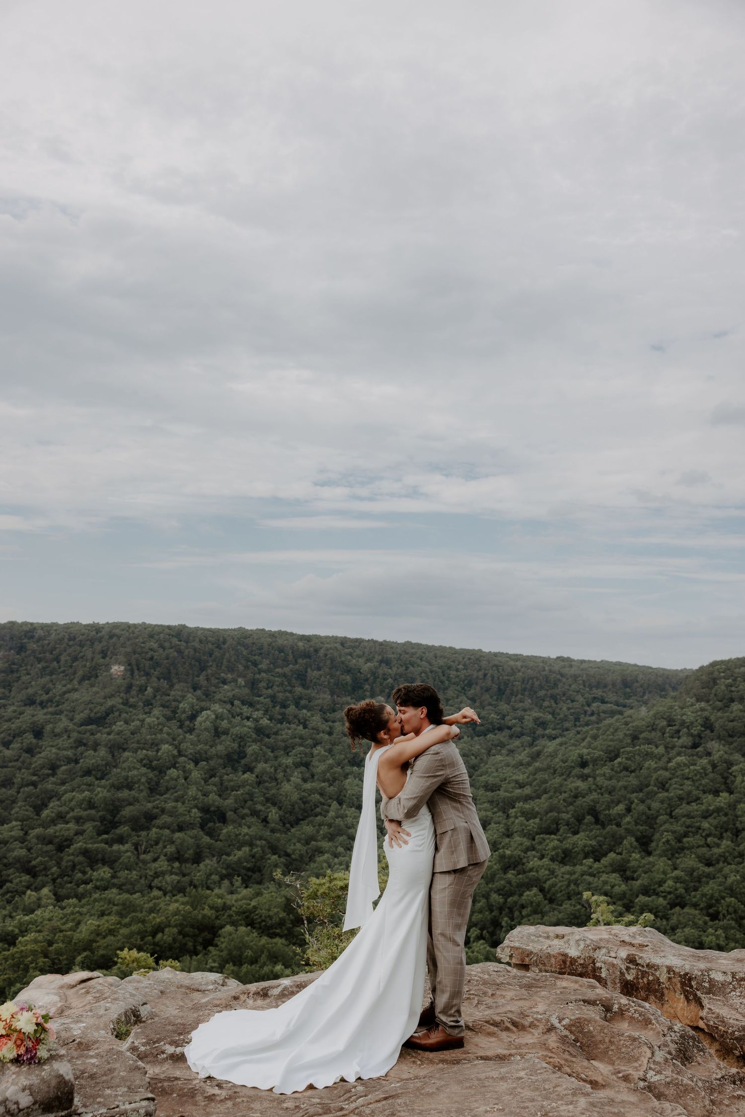 Bride and groom share a kiss on a rocky overlook with lush green forest and cloudy sky backdrop.