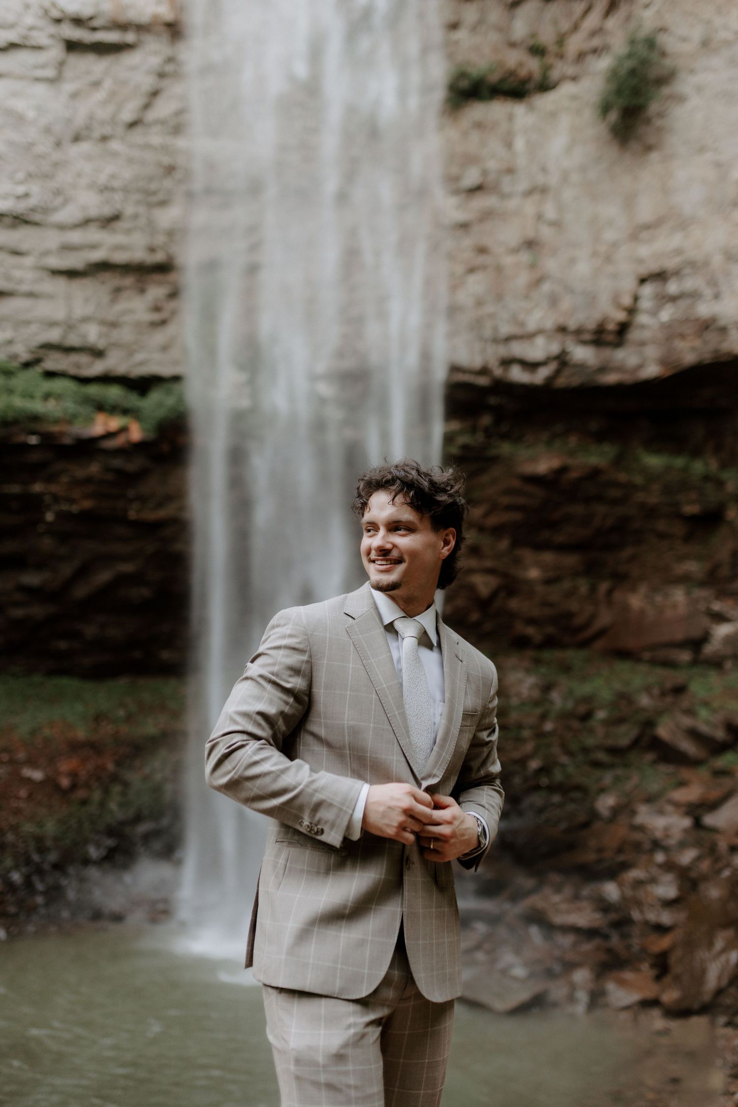 Smiling groom in tan plaid suit stands confidently before a stunning cascading waterfall backdrop.