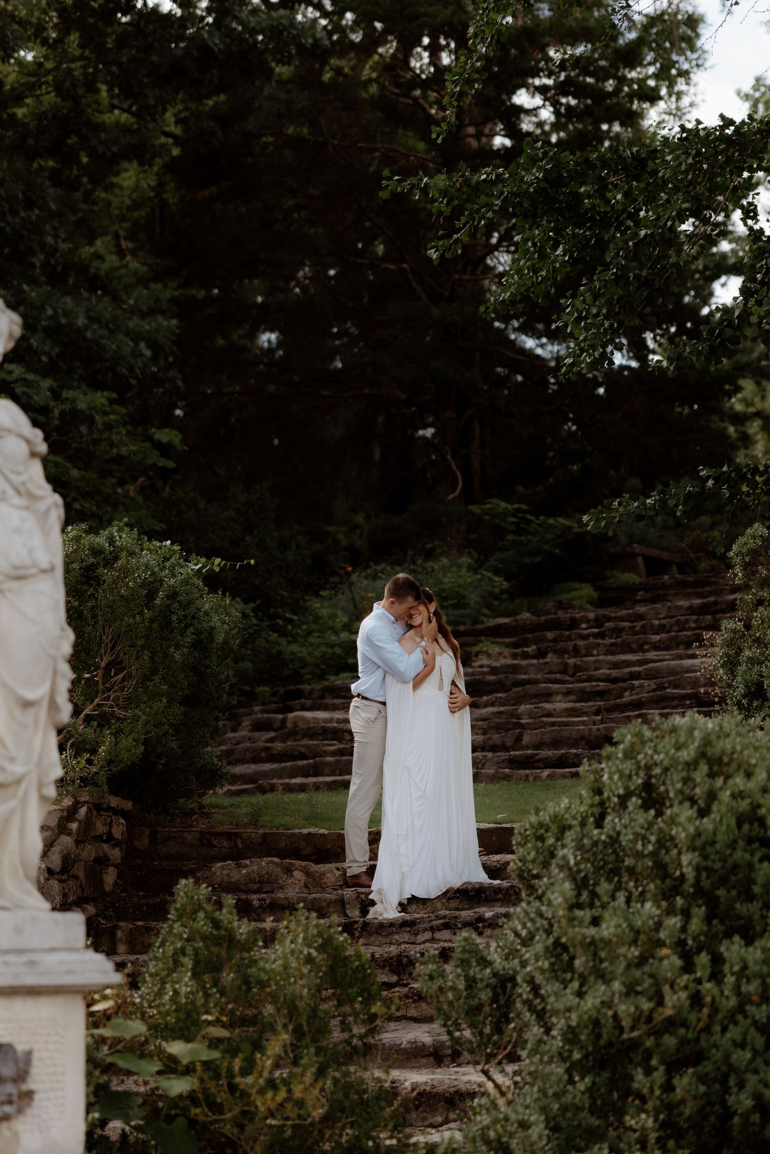 A couple embraces on stone steps surrounded by lush greenery and a classical statue in an elegant garden setting.