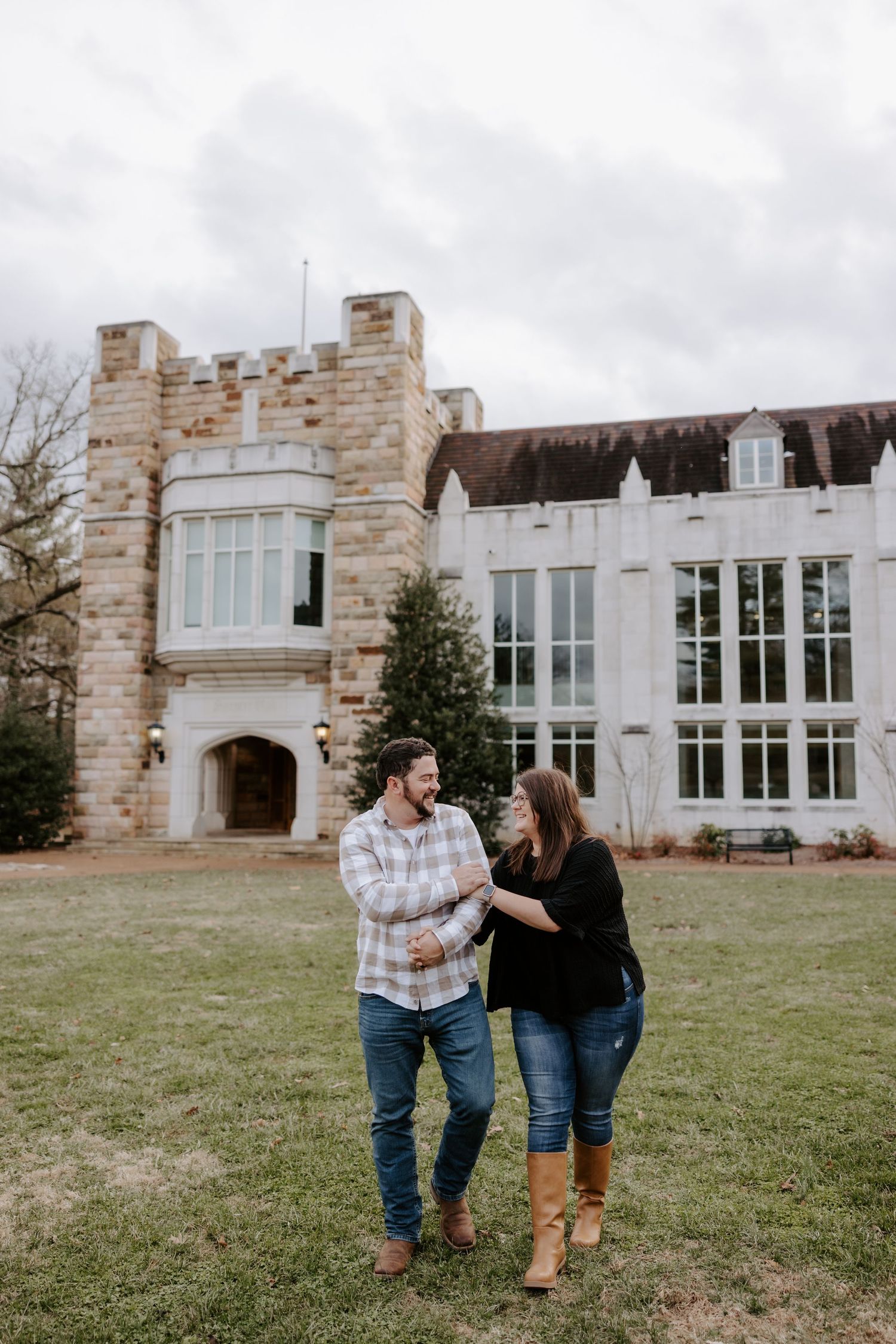 Couple smiling and holding hands in front of a stone castle building on a green lawn under cloudy skies.