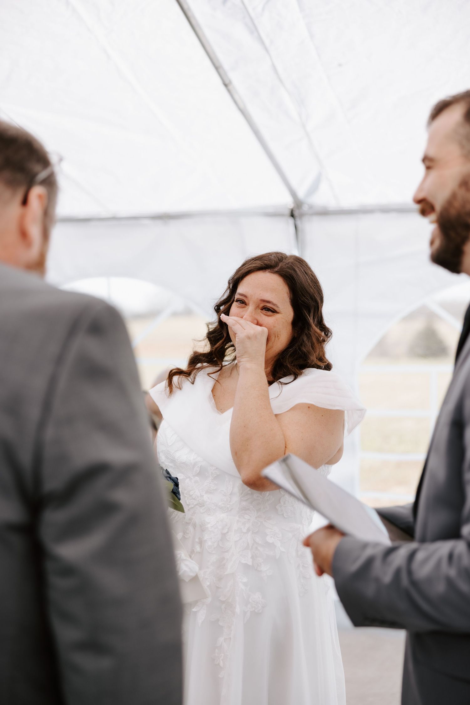 Emotional bride in white dress laughing during outdoor wedding ceremony under tent with groom in gray suit.