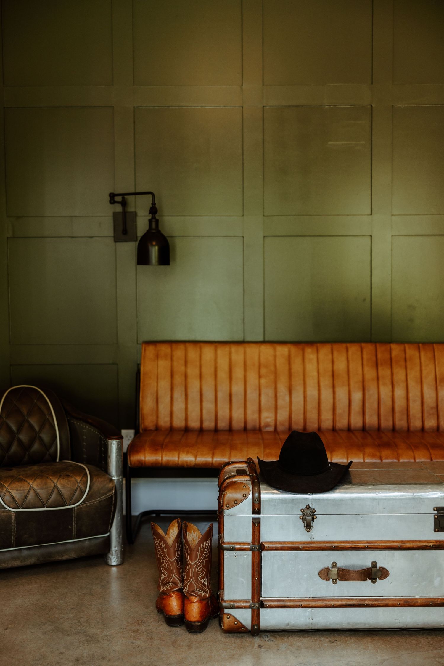 Vintage leather sofa with cowboy boots, hat, and trunk in a rustic green paneled room.