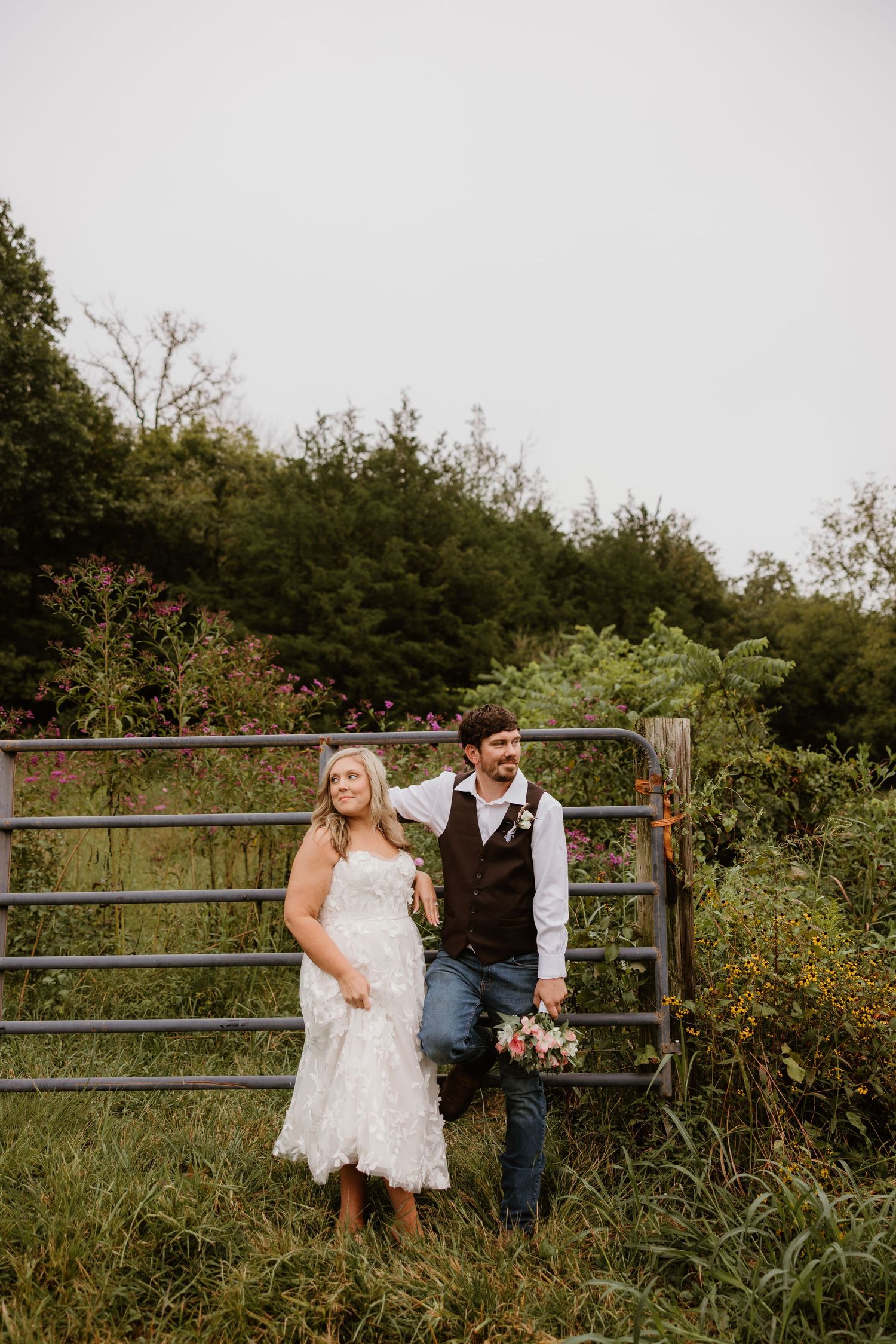 A couple sits on a rustic wooden fence surrounded by lush greenery and wildflowers at sunset.