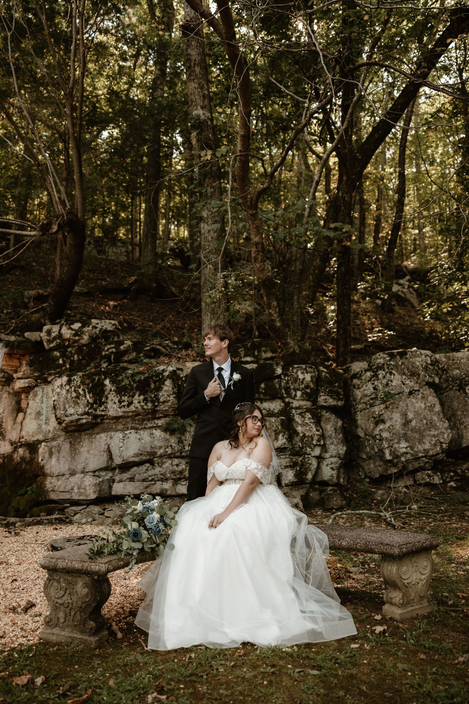 A romantic wedding couple shares intimate moments in a secluded woodland setting with natural rock formations.