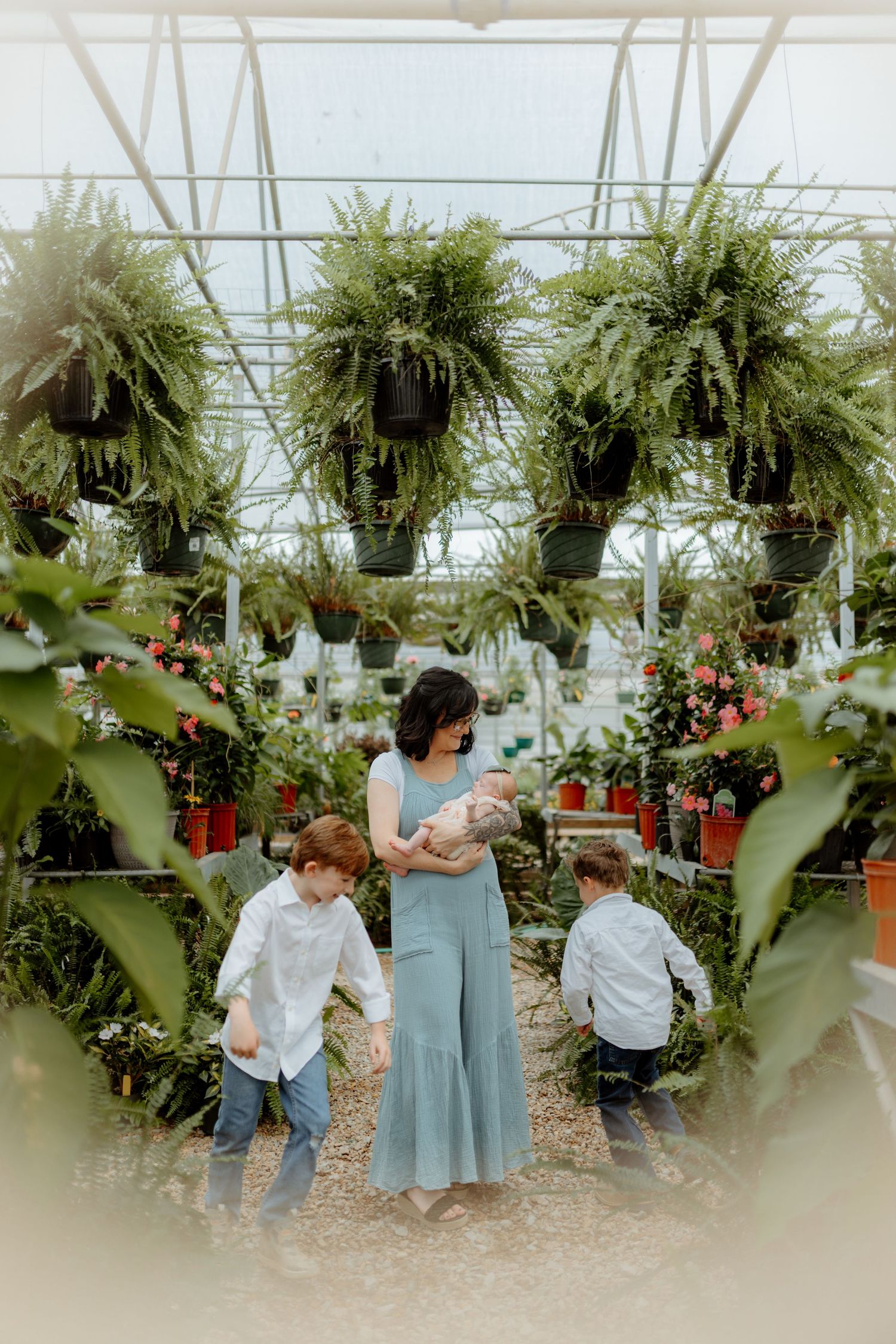 A mother in a blue dress holds her baby while two young boys play around her in a lush greenhouse.
