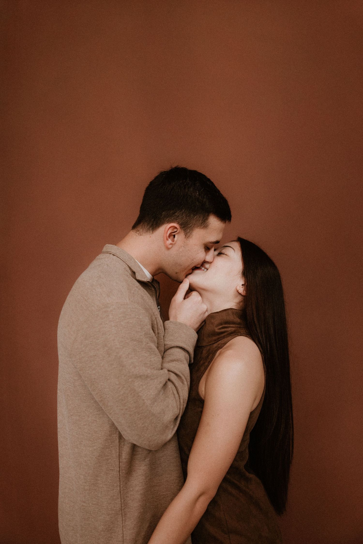 A couple shares a tender almost-kiss moment, dressed in brown tones against a warm terracotta wall.