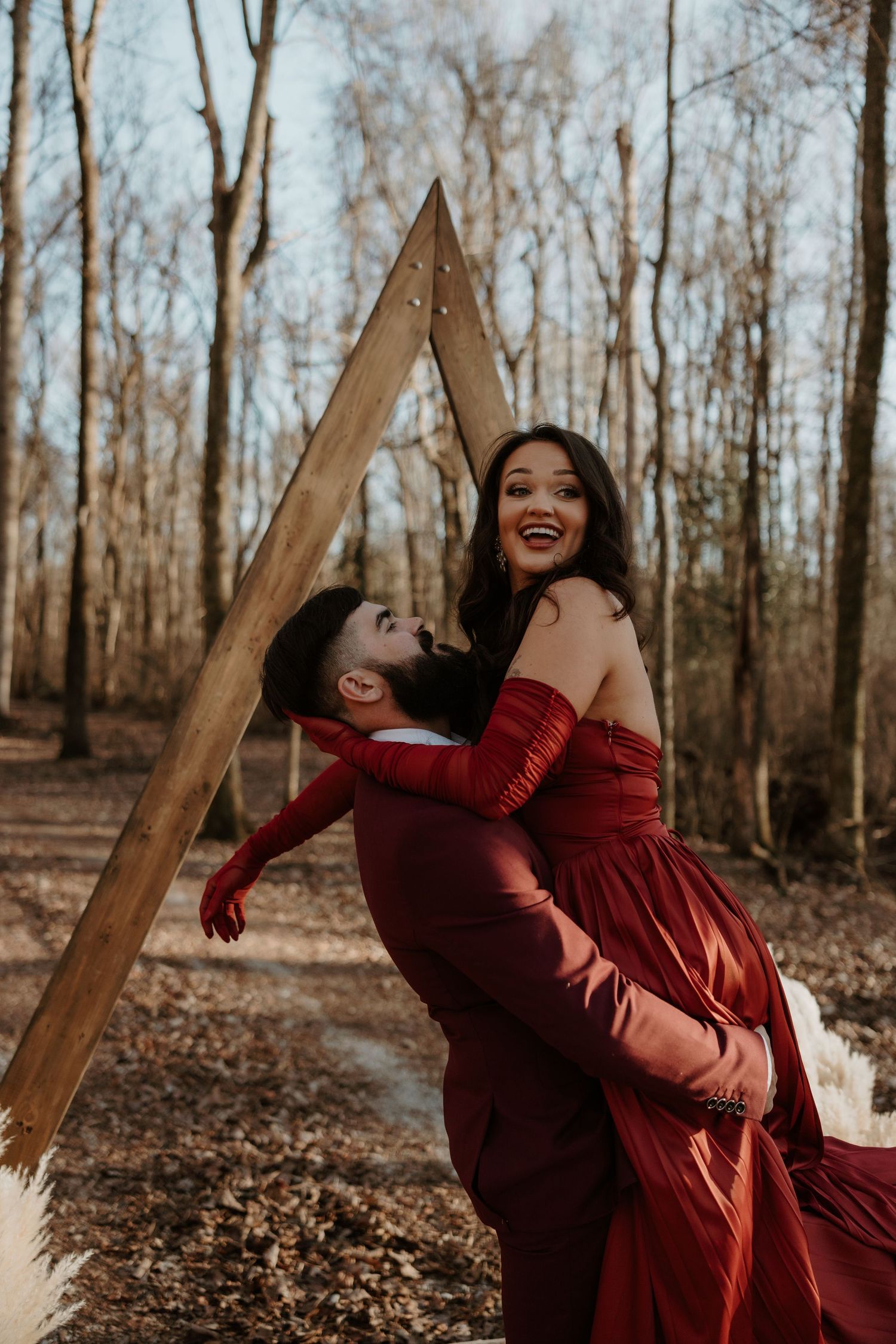 Couple in matching red outfits embracing joyfully in a forest with a wooden triangle arch backdrop.