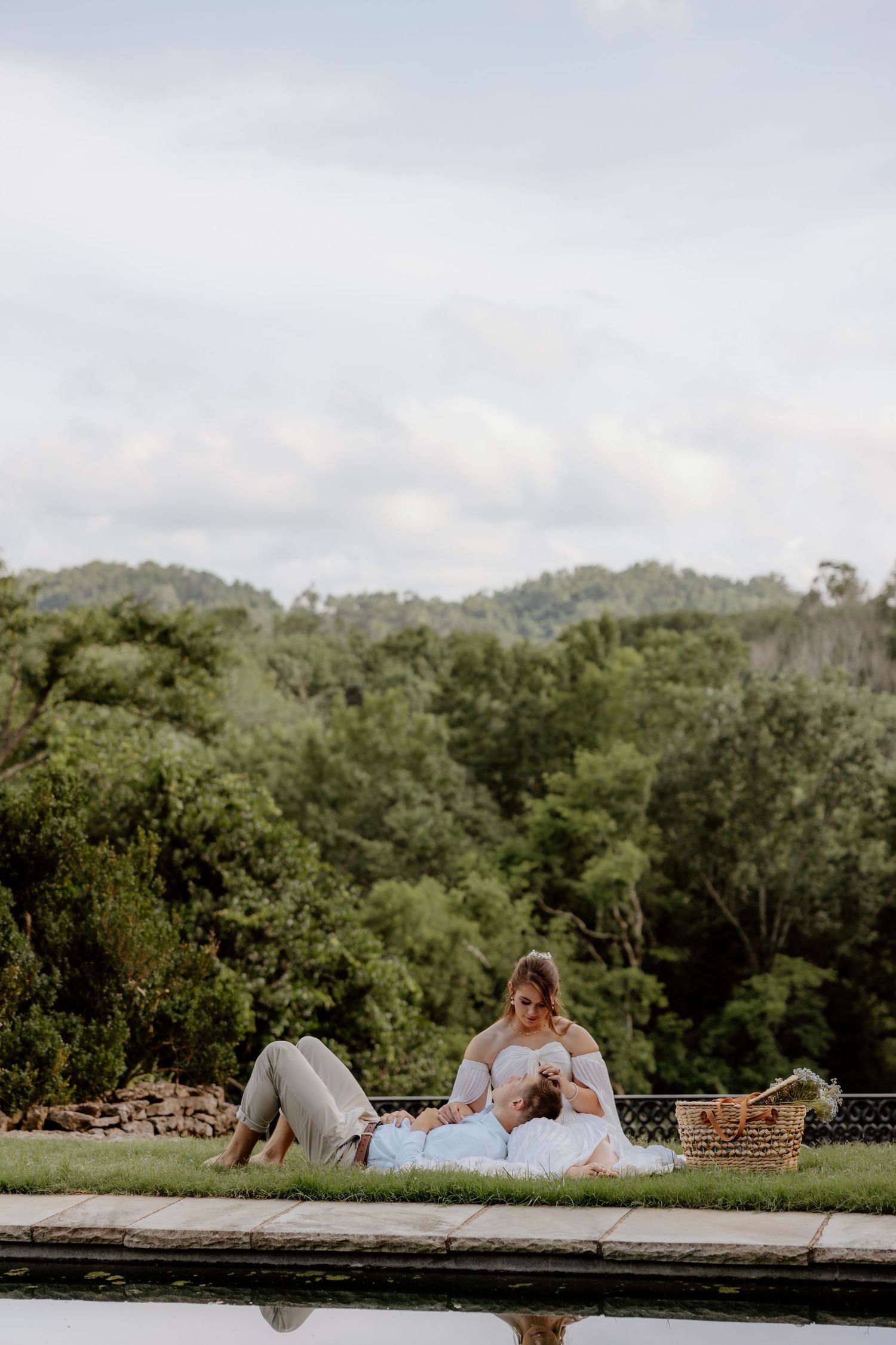 Couple enjoying a romantic picnic by a pool surrounded by lush green trees on a cloudy day.