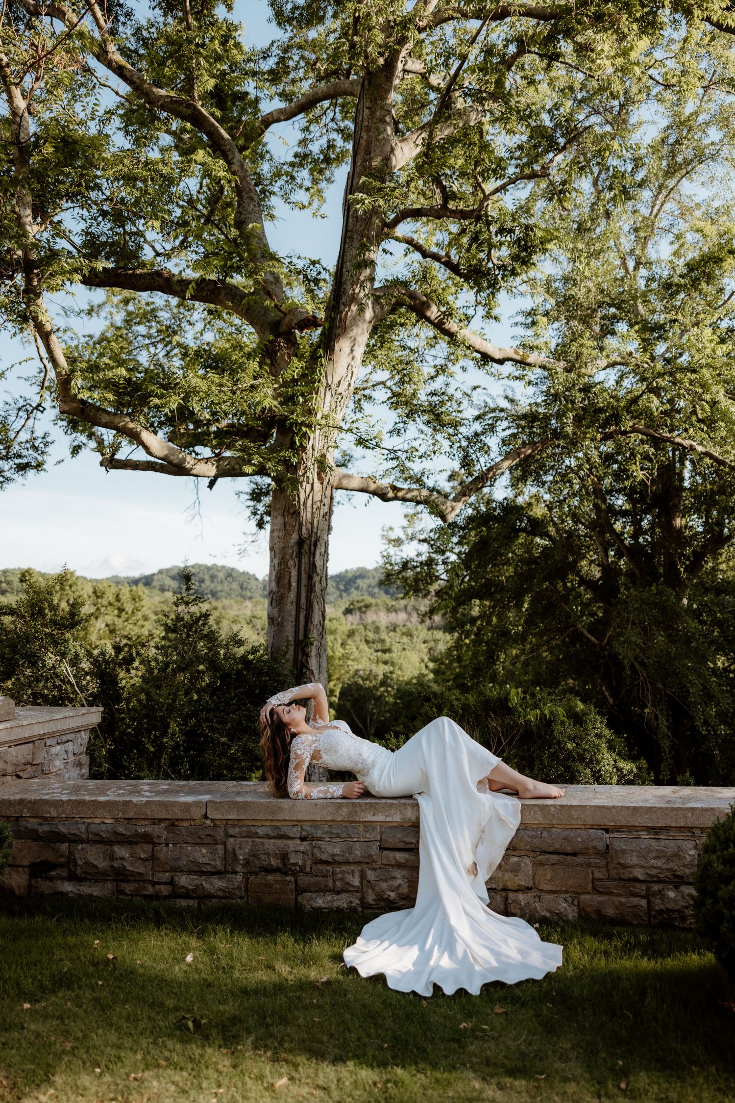 Bride in white gown poses dramatically on stone wall surrounded by lush green trees in outdoor wedding photo.