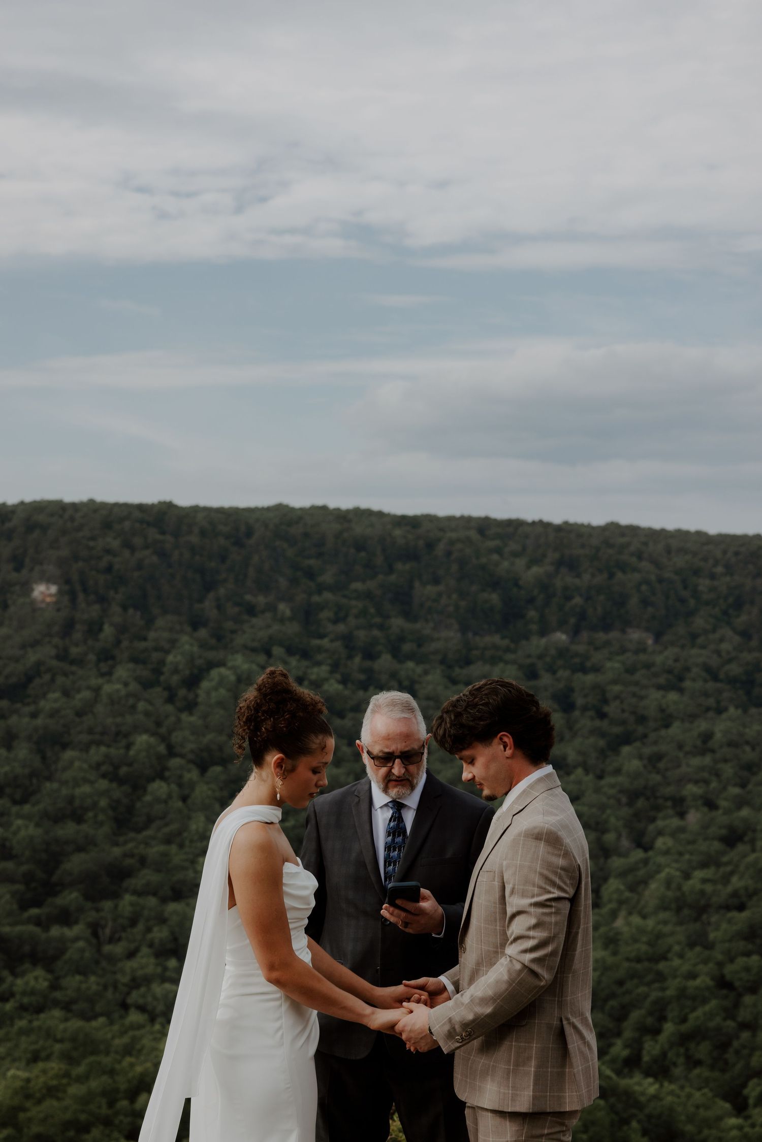 Couple exchanges vows with officiant at an outdoor wedding ceremony overlooking a lush forested hillside.