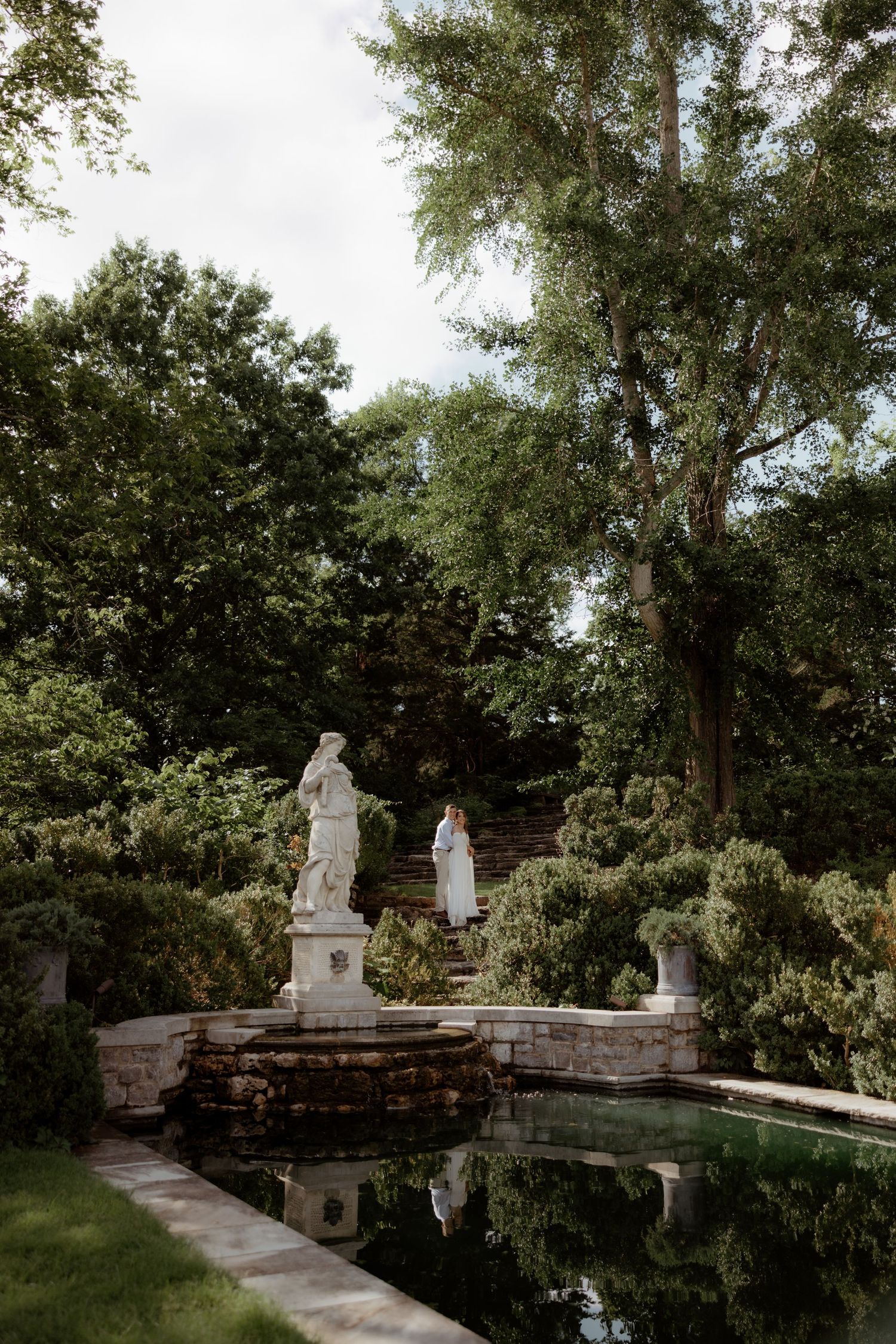 Elegant garden with classical white statues beside a reflective pool, surrounded by lush green trees and stone steps.