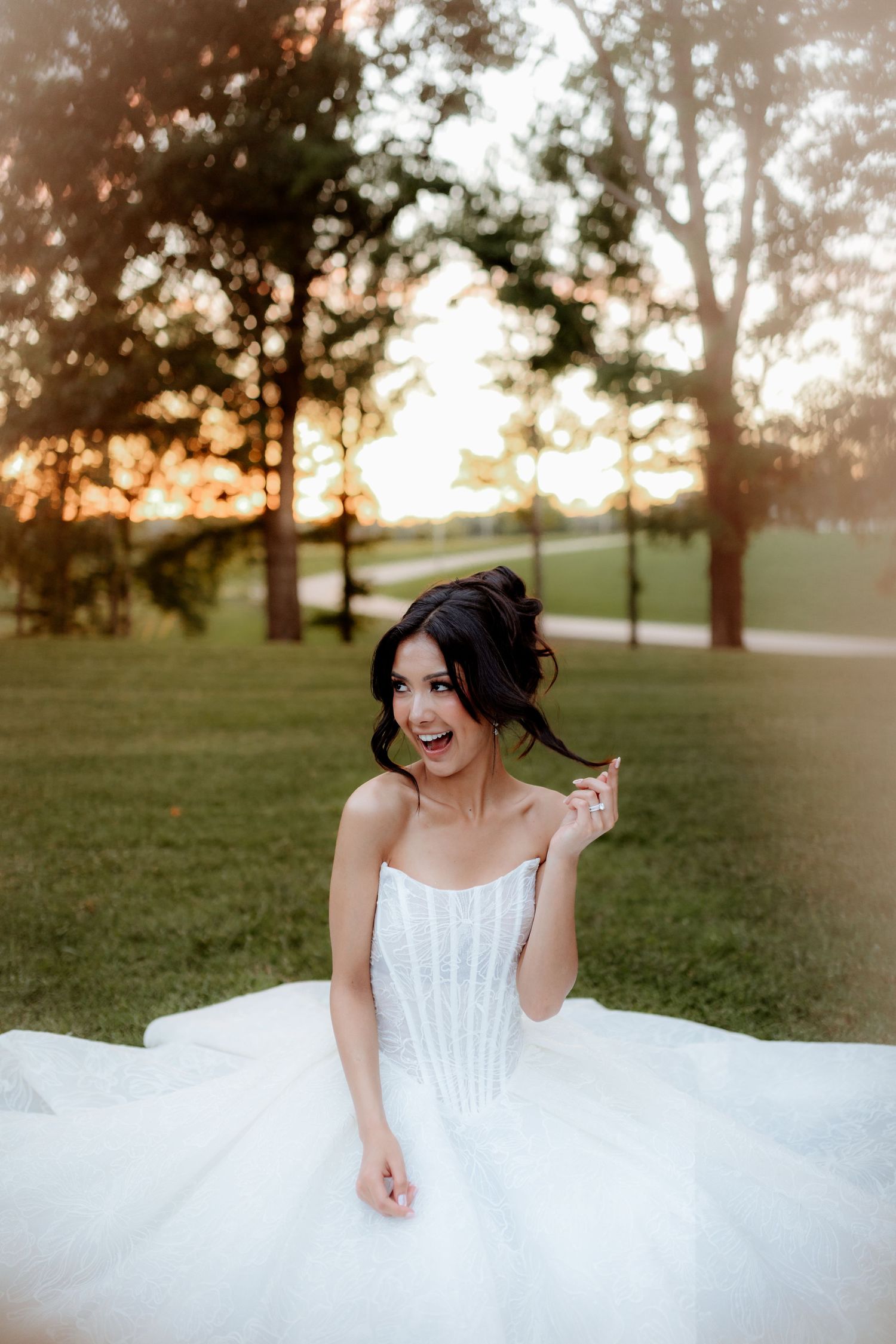Bride in white ballgown sitting on green grass at sunset, smiling joyfully in an outdoor park setting.