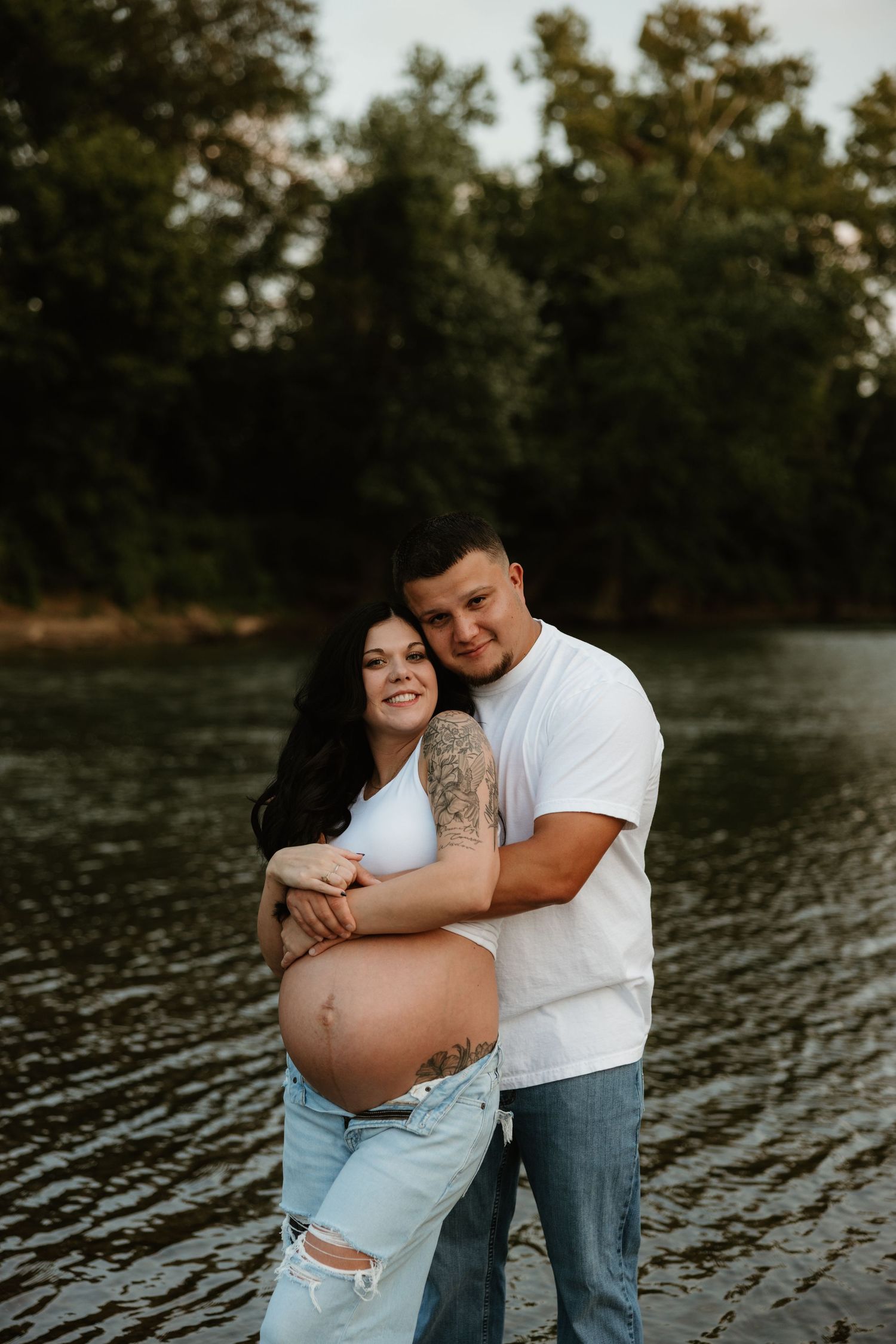 A romantic maternity photoshoot by the water features a couple sharing an intimate moment at sunset.