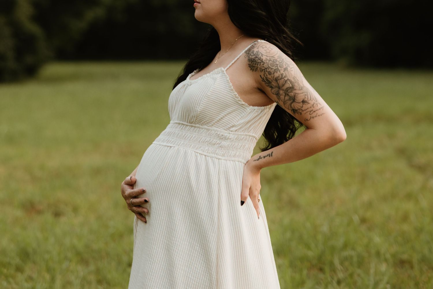 A pregnant woman in a white sundress poses outdoors in a grassy field at golden hour.