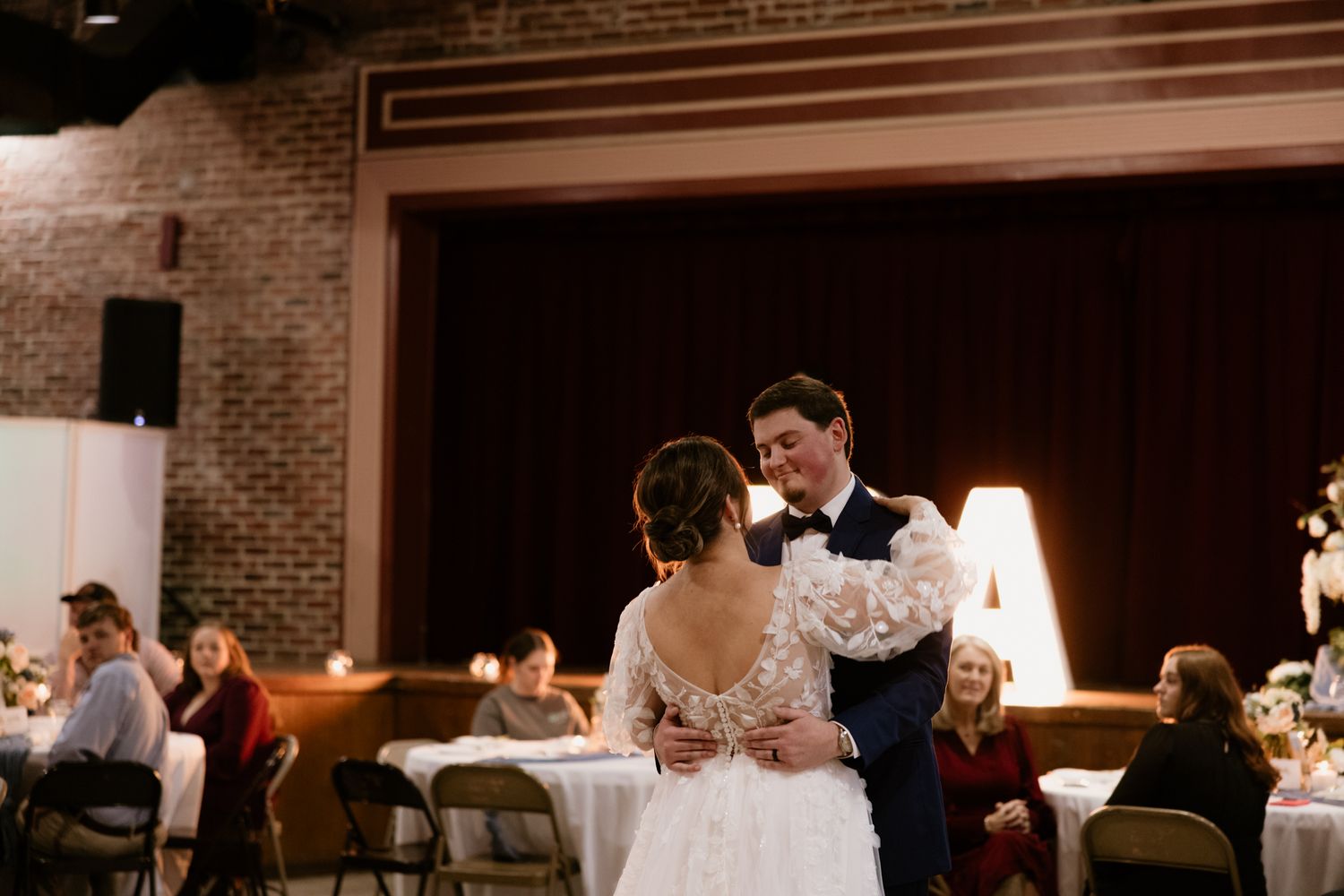 A couple shares their first dance in an elegantly lit reception hall with brick walls.