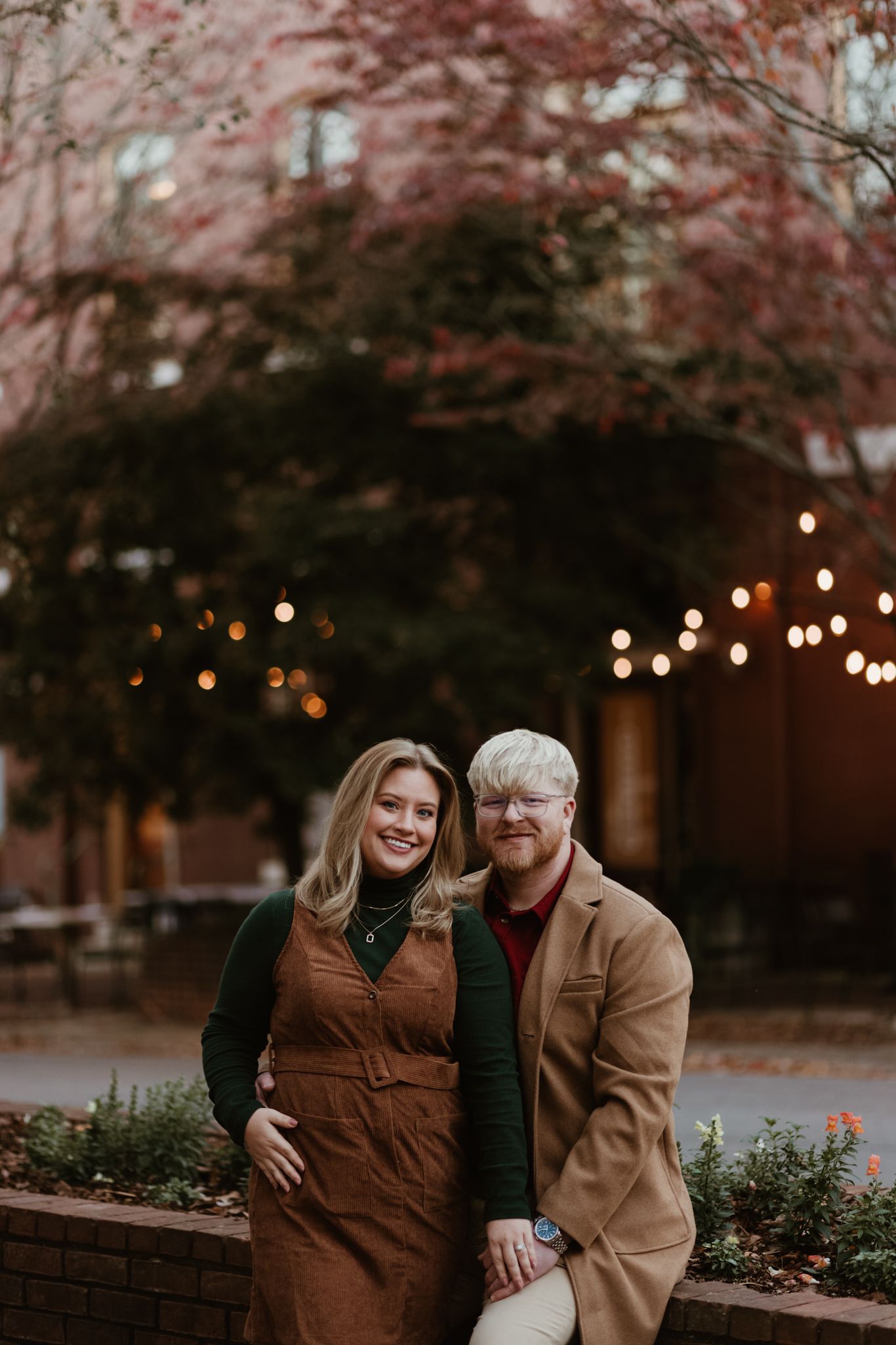 A couple poses together outdoors at dusk with string lights and autumn foliage in the background.