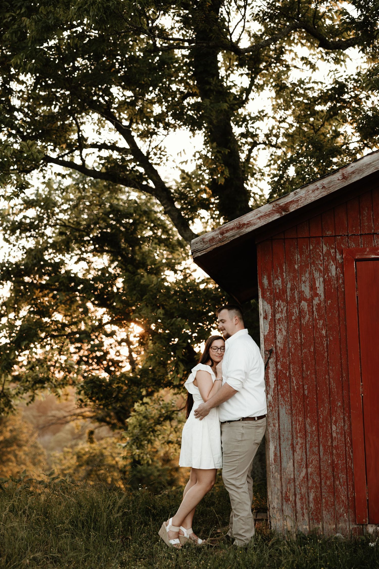 A couple embraces outside a rustic red barn at sunset with green trees in the background.