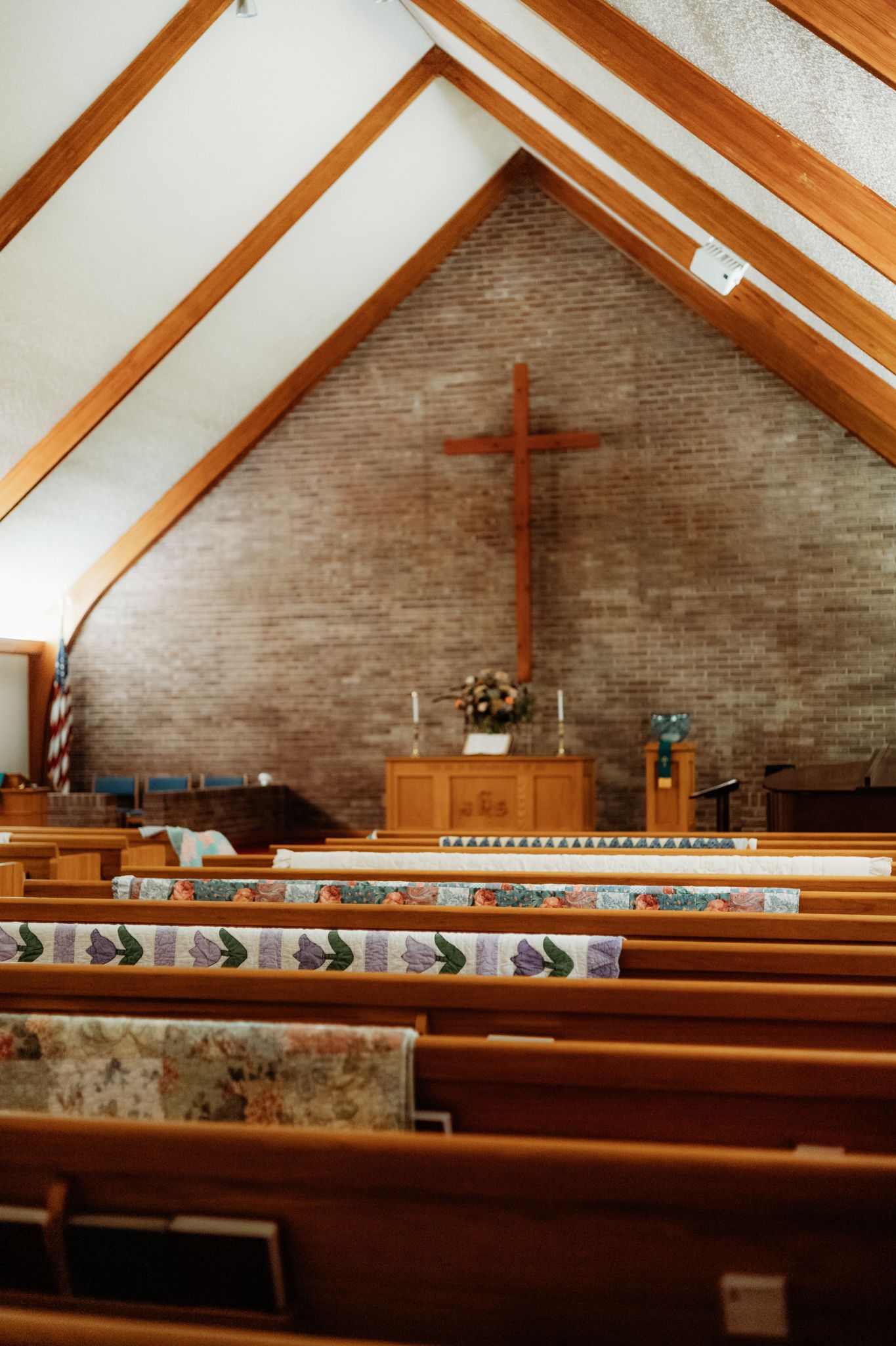 Interior of a modern church with wooden beams, pews, and a simple cross mounted on a brick wall in warm lighting.