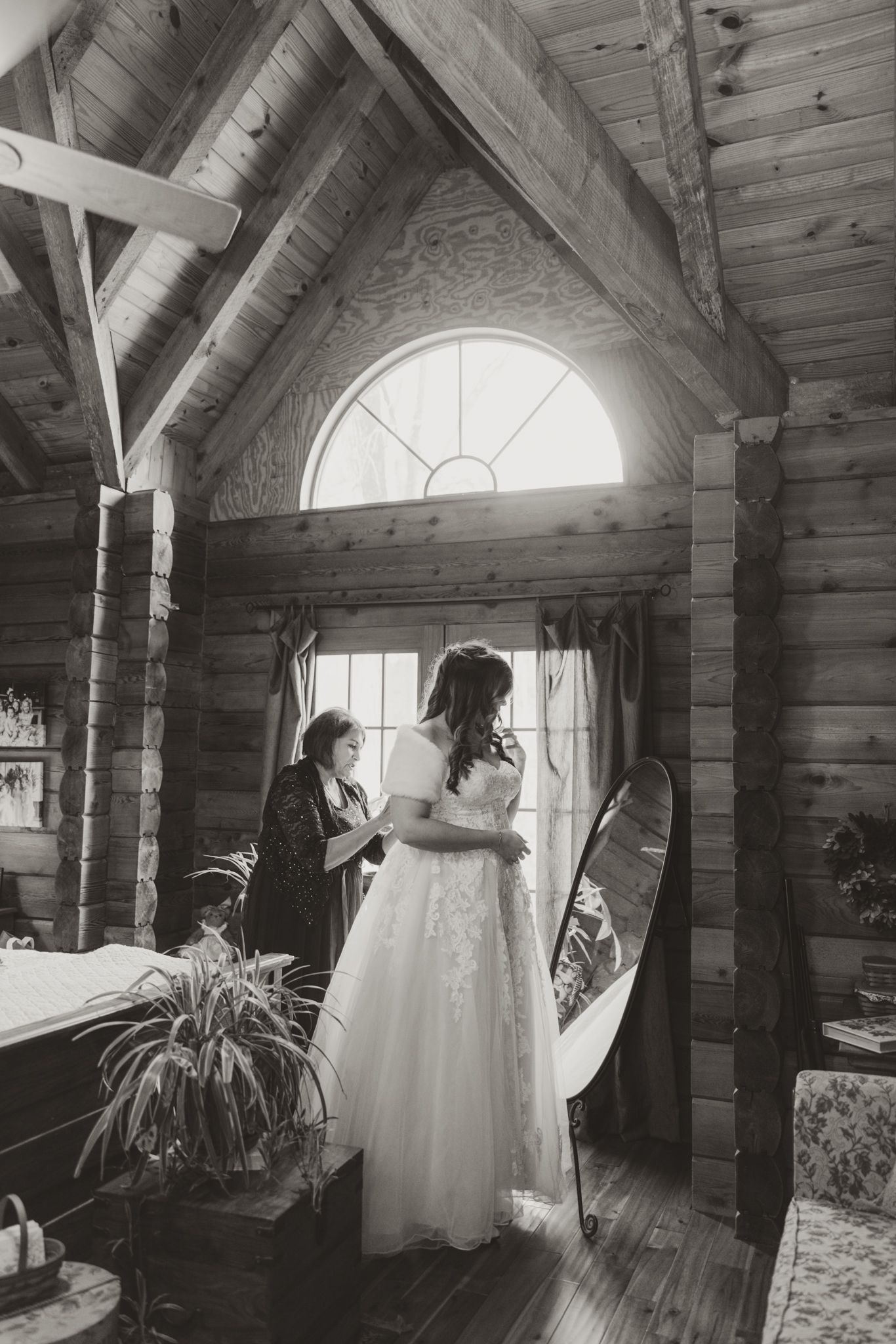 Black and white photo of a rustic log cabin interior with a wedding dress and full-length mirror by an arched window.