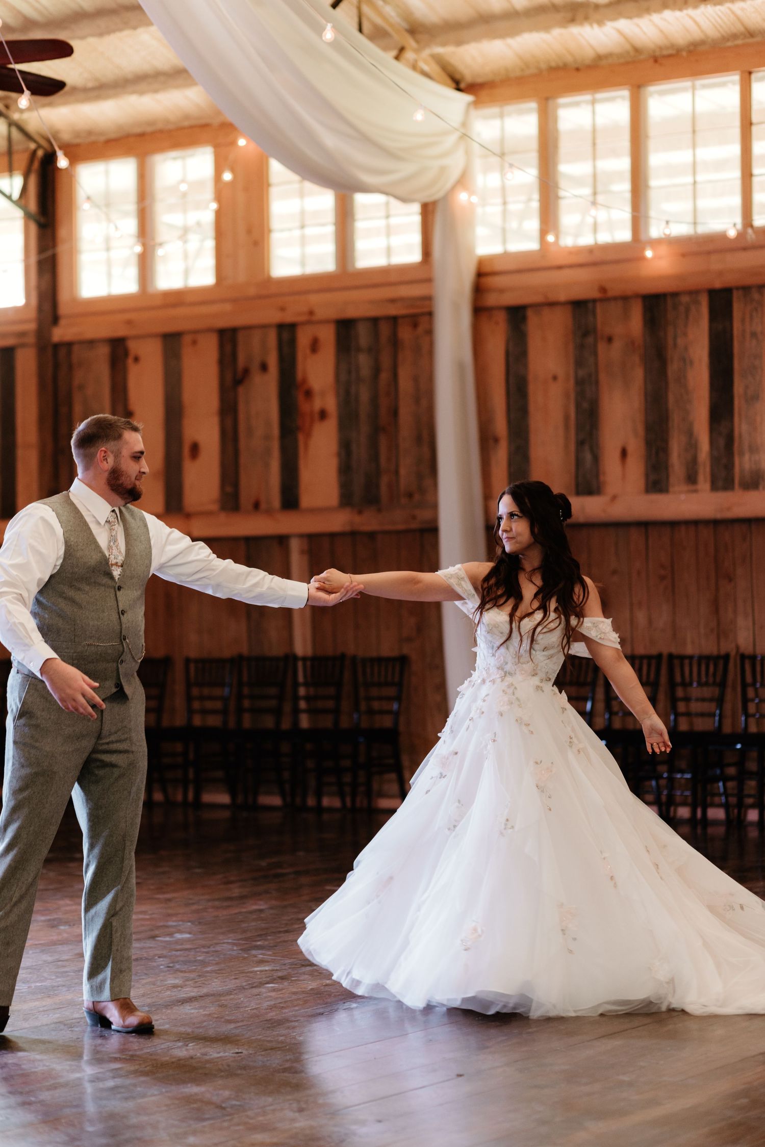 A couple dancing in a rustic barn venue with draped white fabric and natural light streaming through windows.