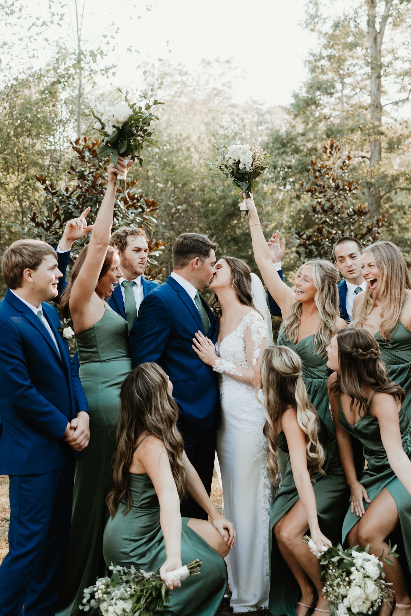Wedding party celebrating outdoors with raised bouquets while wearing navy suits and sage green dresses.