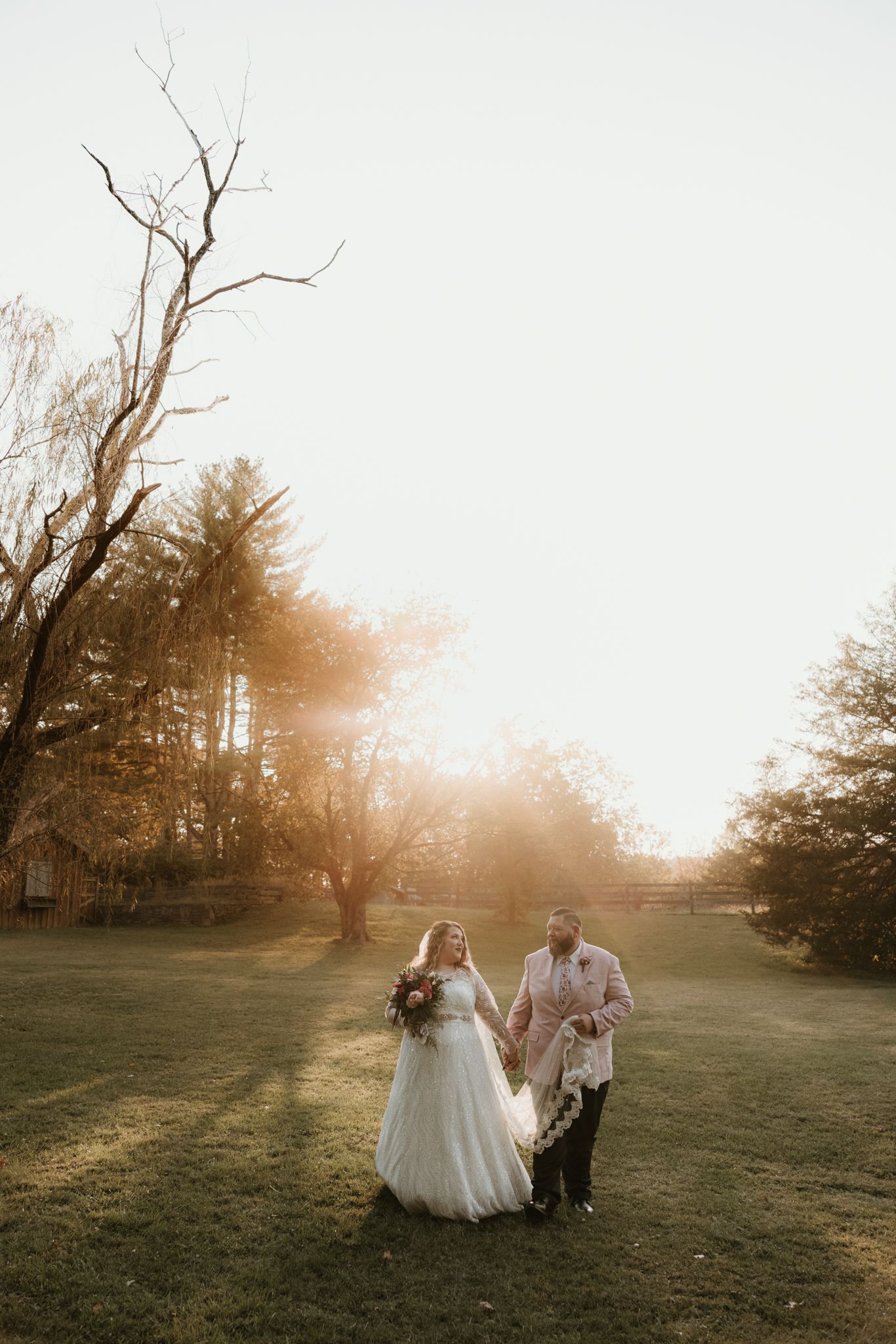 Golden sunlight bathes a grassy field as elegant figures in wedding attire walk together during a sunset photoshoot.