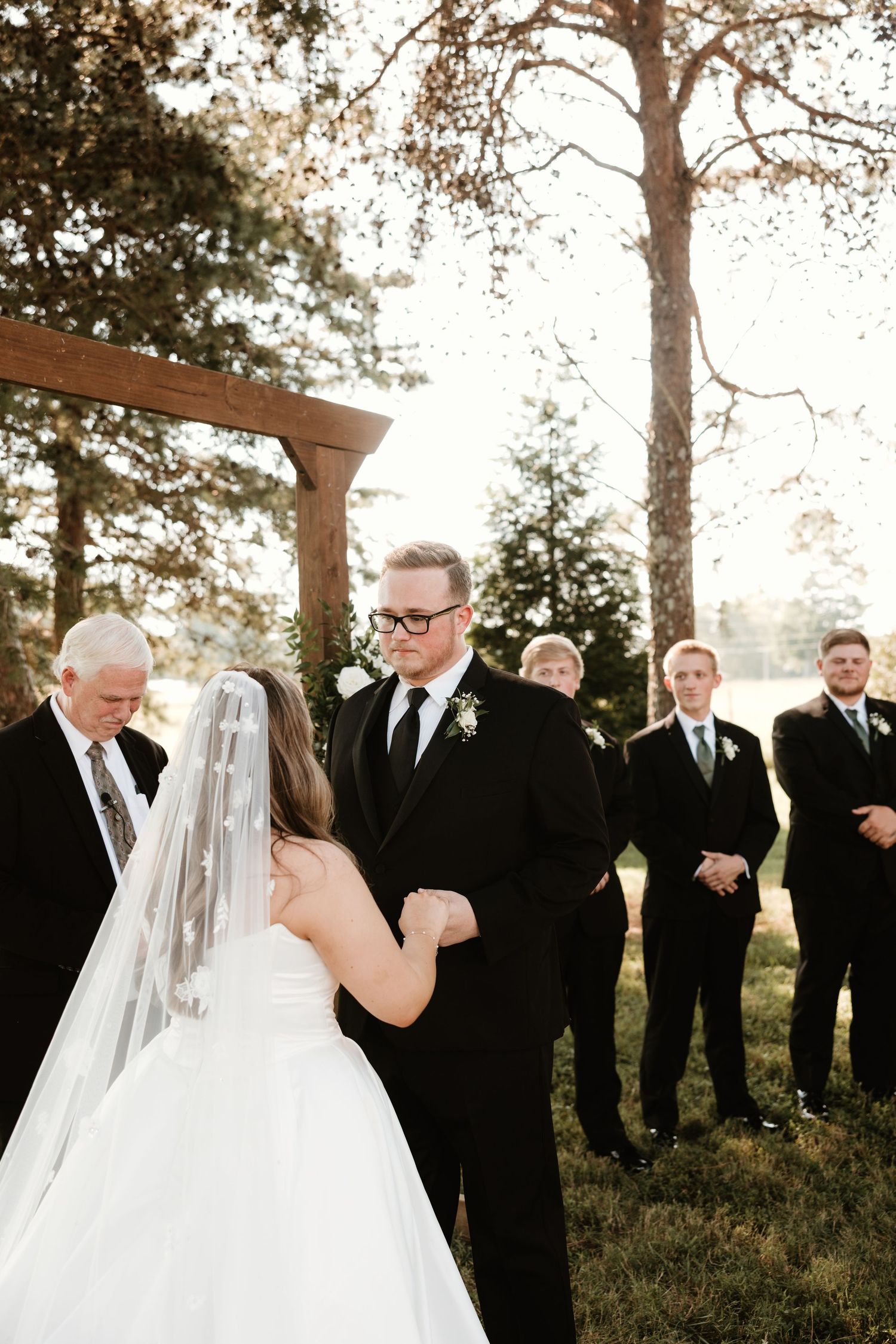 Wedding sequence shows a bride and groomsmen lined up outdoors under tall evergreen trees at sunset.